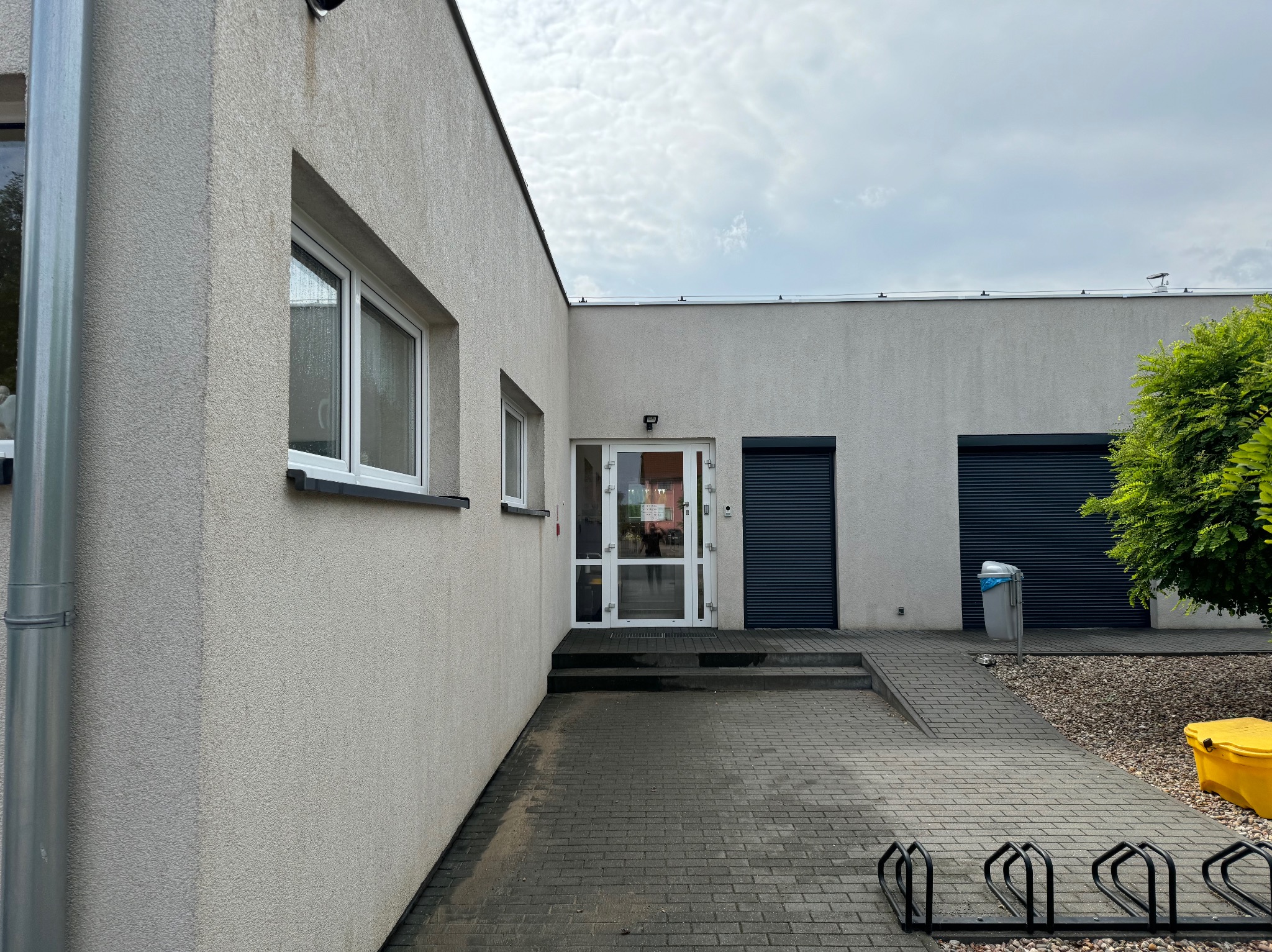 Budynek z jasną elewacją, widoczne okna, drzwi wejściowe, rynna, fragment chodnika z kostki brukowej, stojak na rowery, kosz na śmieci i fragment zieleni.
