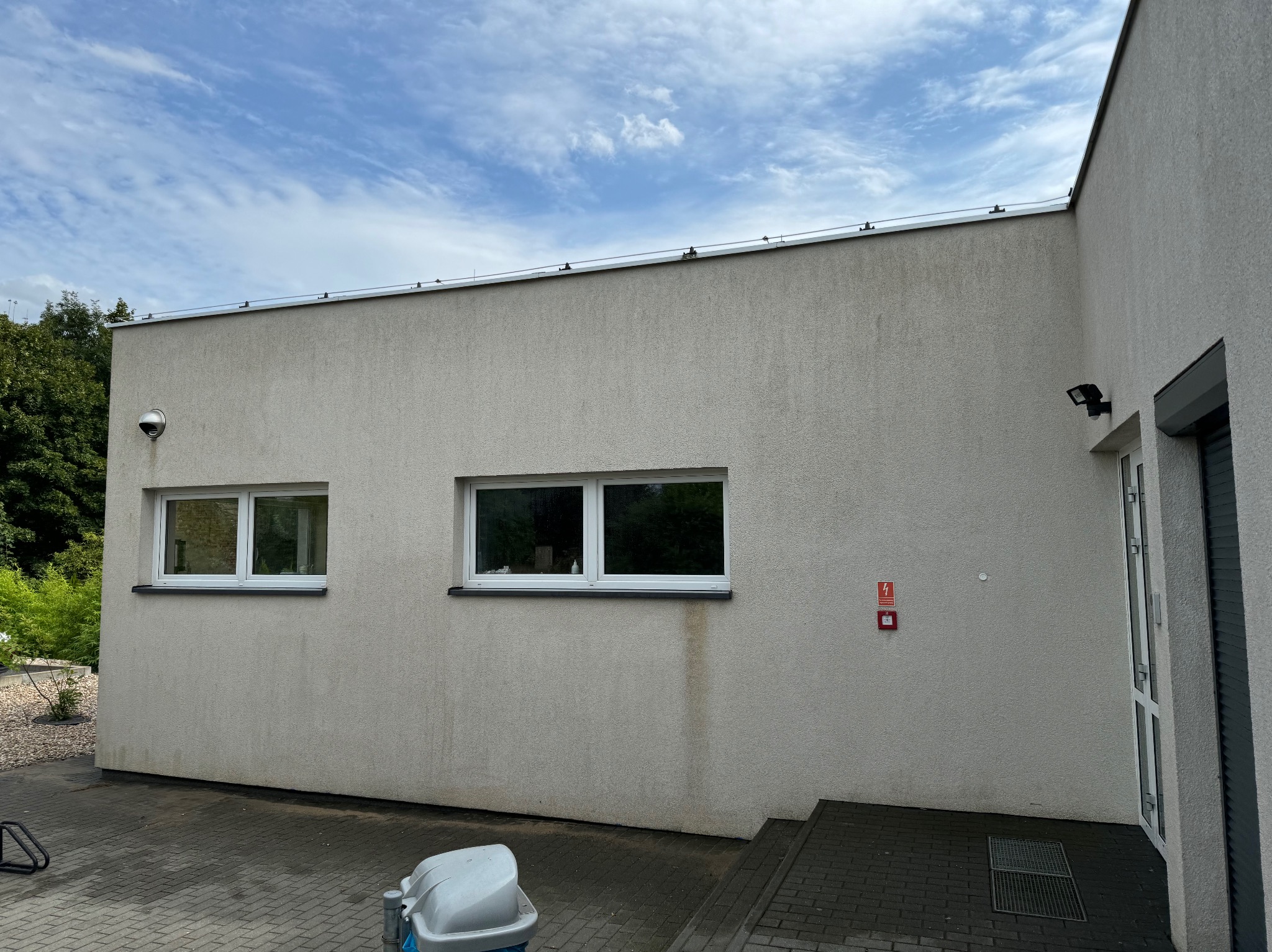 Jasna elewacja budynku z dwoma oknami i widocznymi śladami zacieków poniżej jednego z nich, alarm przeciwpożarowy na ścianie, kostka brukowa na dziedzińcu