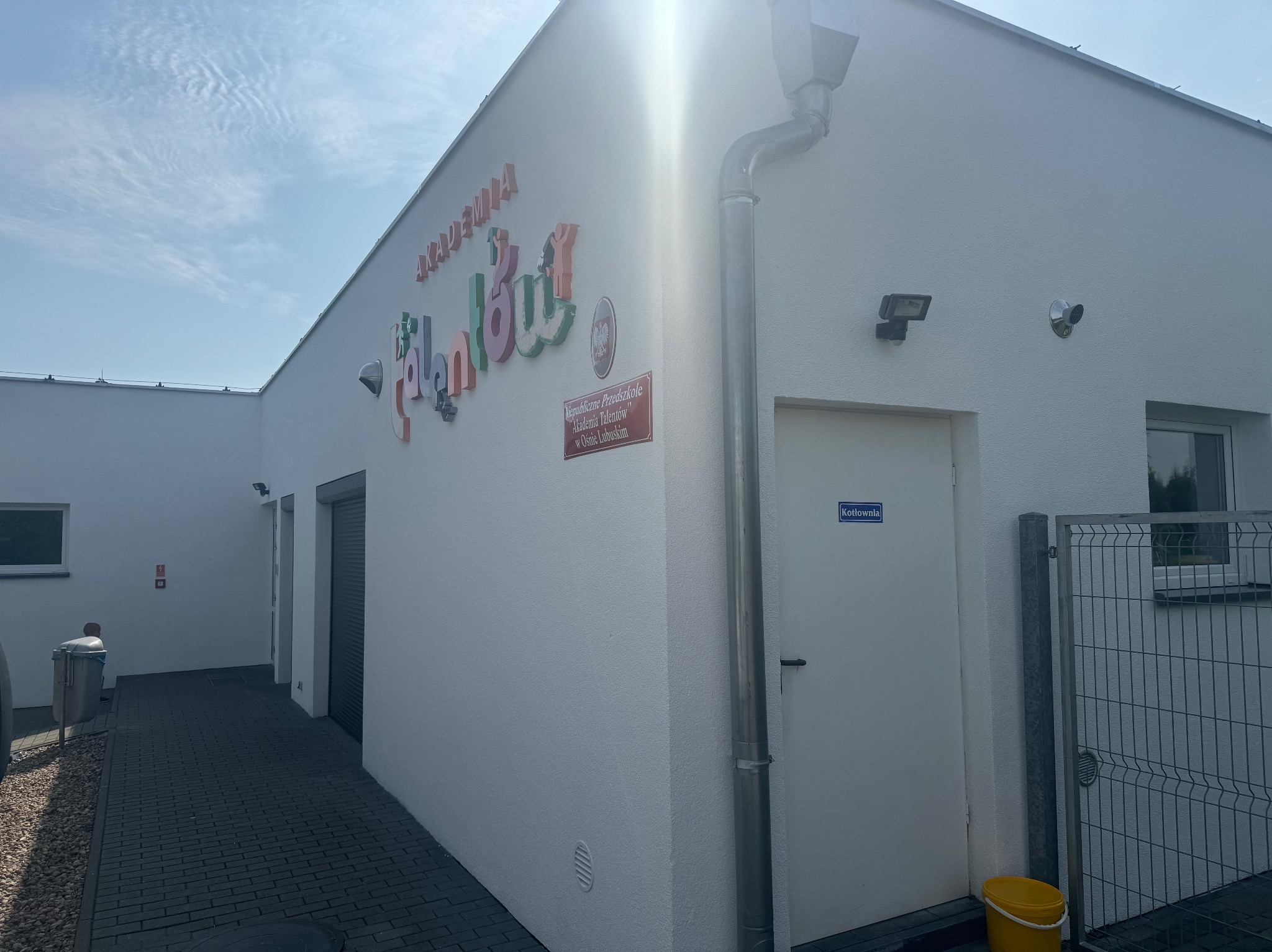 Biały budynek przedszkola z kolorowym napisem 'Akademia Talentów', widoczne rynny, lampy zewnętrzne i kostka brukowa przed budynkiem.