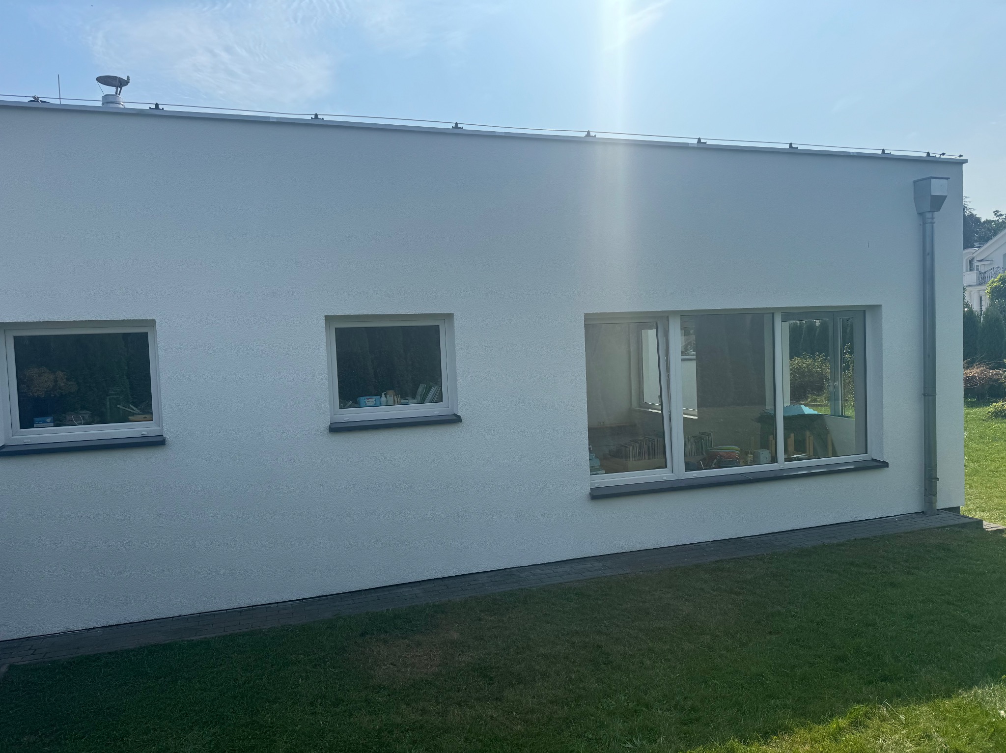 Boczny widok białej elewacji domu jednorodzinnego z trzema oknami, widoczne rynna i trawnik przed budynkiem w słoneczny dzień.