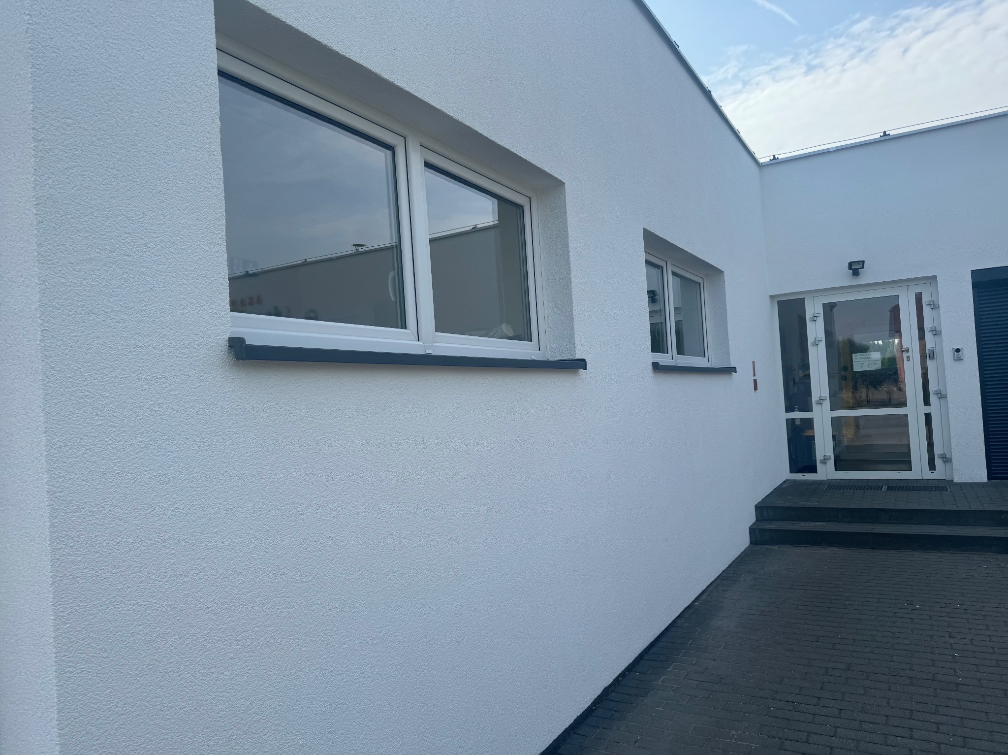 Świeżo pomalowana, biała elewacja budynku z dwoma oknami i wejściem, widoczna faktura tynku baranka.