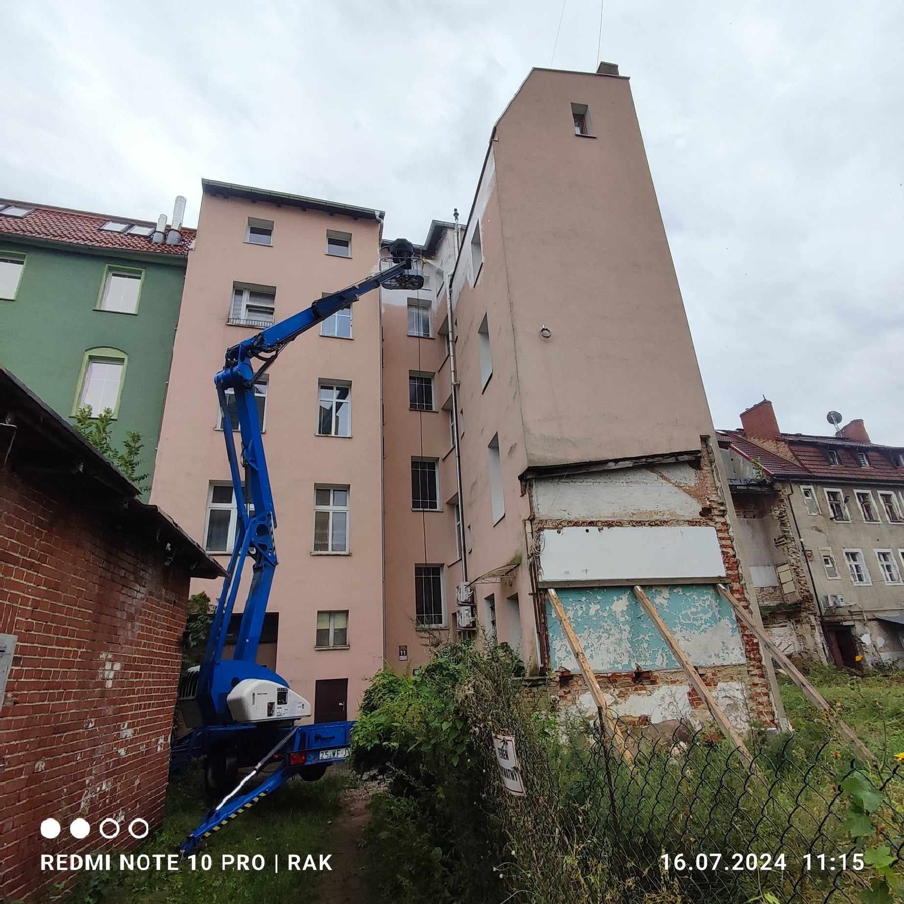 Praca na wysokościach: niebieski podnośnik koszowy przy elewacji budynku w trakcie malowania, widoczne ślady świeżej farby, częściowo odnowiona fasada, otoczenie zaniedbane z bujną roślinnością...