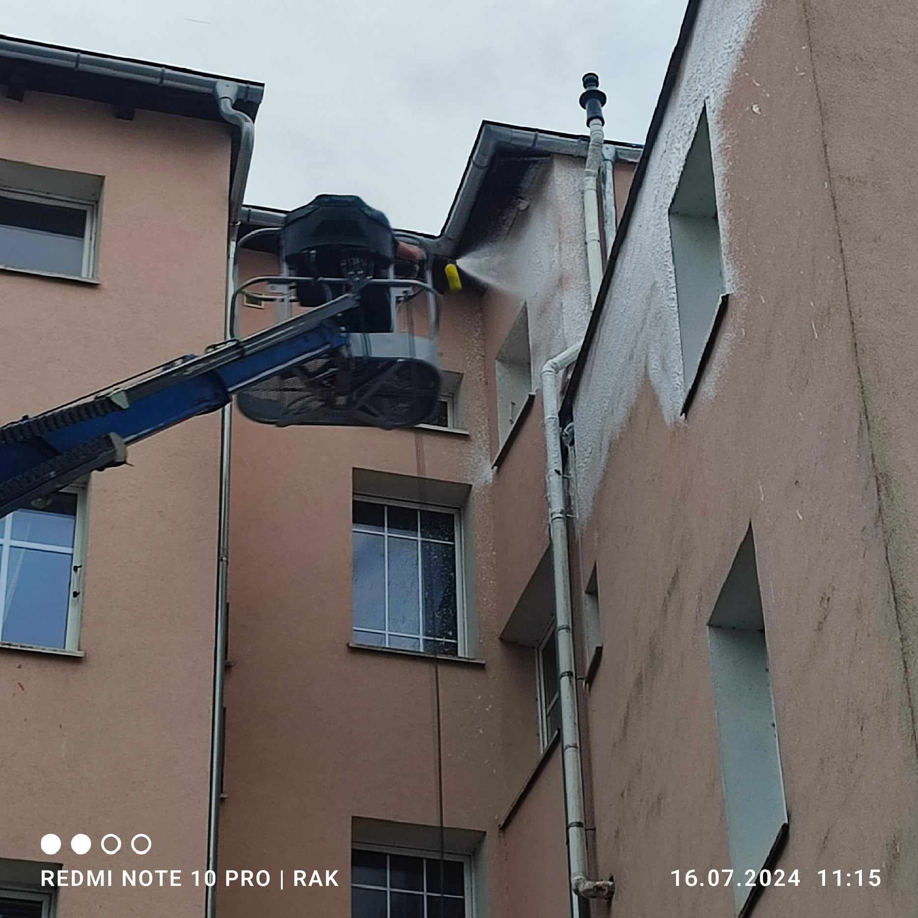 Pracownik na podnośniku koszowym spryskuje elewację budynku strumieniem wody pod ciśnieniem, czyszcząc ją z zabrudzeń. Widoczne ślady spływającej wody i różnica w czystości powierzchni.