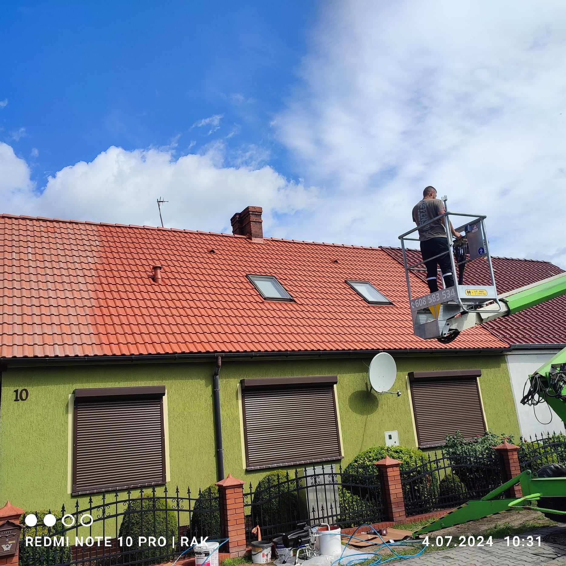 Pracownik na podnośniku koszowym w trakcie czyszczenia połowy dachu z czerwonej dachówki, widoczna różnica w kolorze między czystą a zabrudzoną częścią, zielona elewacja domu z brązowymi roletami...