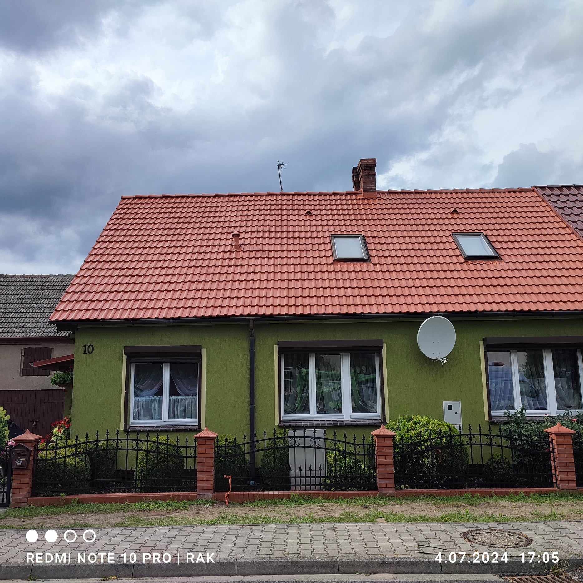 Dom jednorodzinny z czerwoną dachówką, zieloną elewacją, numerem 10 i anteną satelitarną na ścianie, sfotografowany w pochmurny dzień.