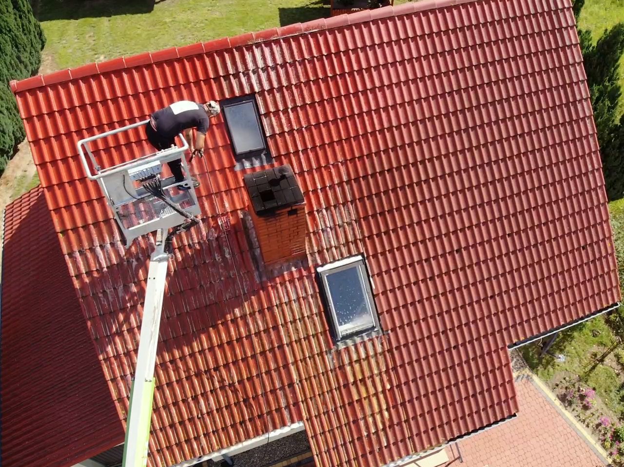 Pracownik myje czerwoną dachówkę z podnośnika koszowego, widoczny komin, okna dachowe i częściowo oczyszczona powierzchnia dachu.