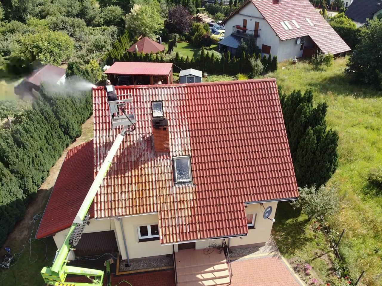 Pracownik na podnośniku koszowym czyści dach z czerwonej dachówki wodą pod ciśnieniem, widoczny efekt czyszczenia - część dachu umyta, część zabrudzona, dom z jasną elewacją w tle.
