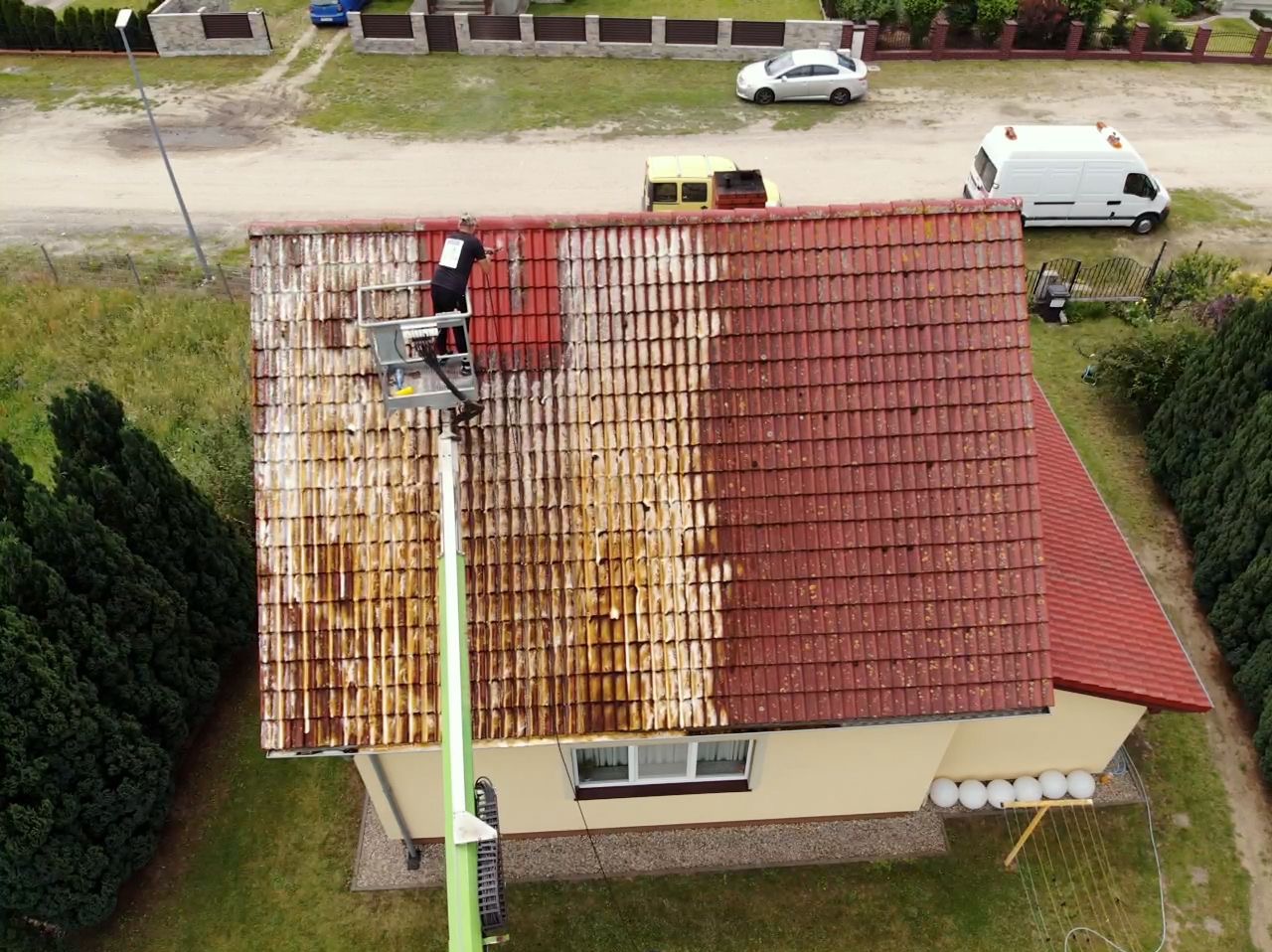 Pracownik na podnośniku koszowym czyści połowę dachu z czerwonej dachówki, widoczny efekt przed i po czyszczeniu, otoczenie to trawnik, dom i droga.