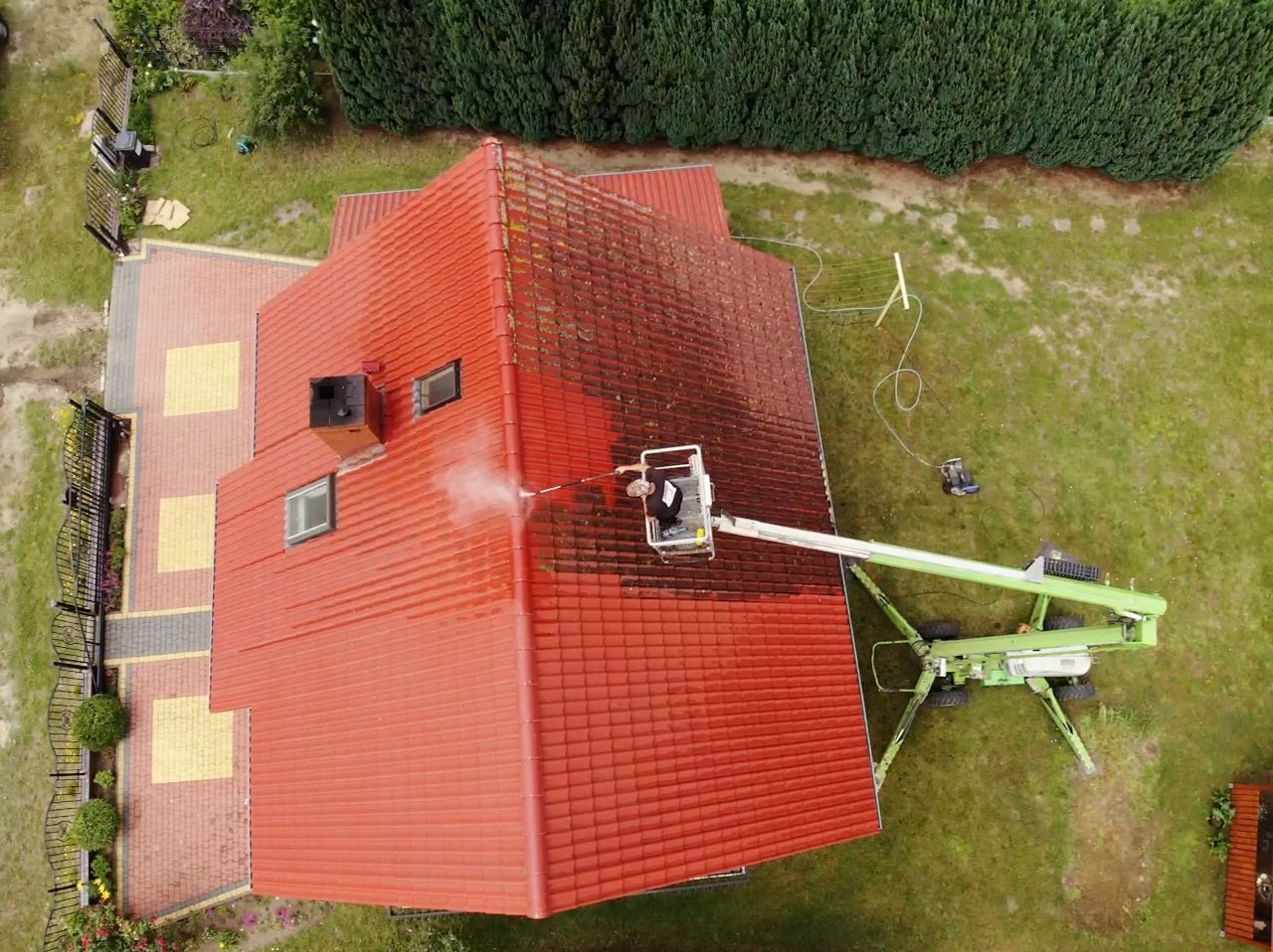 Widok z góry na czerwony dach z dachówki podczas czyszczenia myjką ciśnieniową z podnośnika koszowego; część dachu umyta, druga pokryta mchem i zabrudzeniami.
