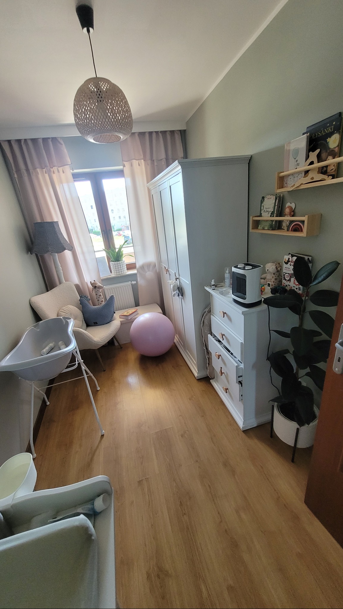 Pokój dziecięcy po odmalowaniu, z nową drewnianą podłogą, jasnymi meblami, lampą wiszącą z plecionki i dekoracjami dla dziecka.