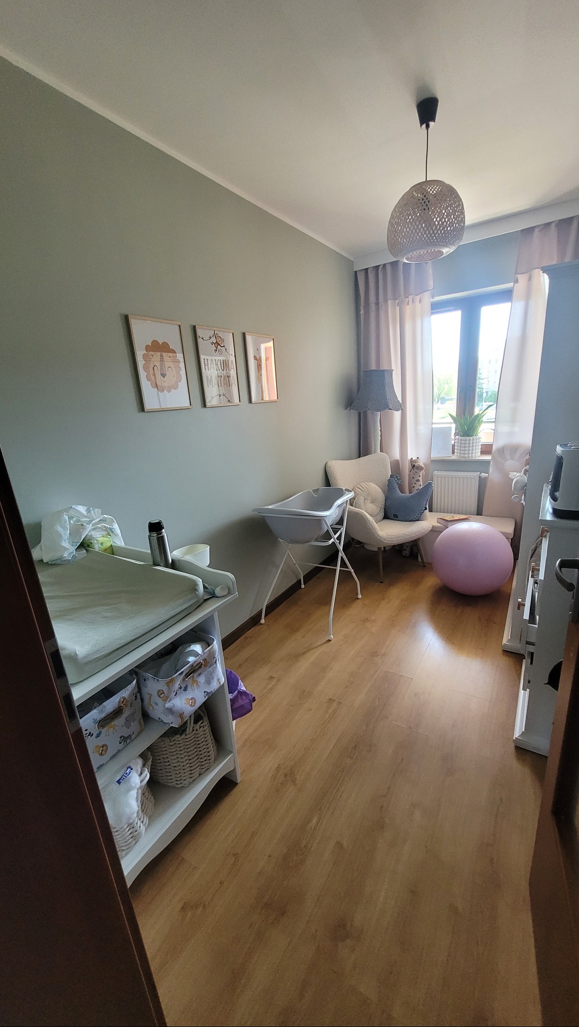 Pokój dziecięcy z drewnianą podłogą, zielonymi ścianami, wiszącą lampą z abażurem, przewijakiem z koszami, wanienką na stojaku, fotelem z poduszką, piłką do ćwiczeń i obrazkami na ścianie.