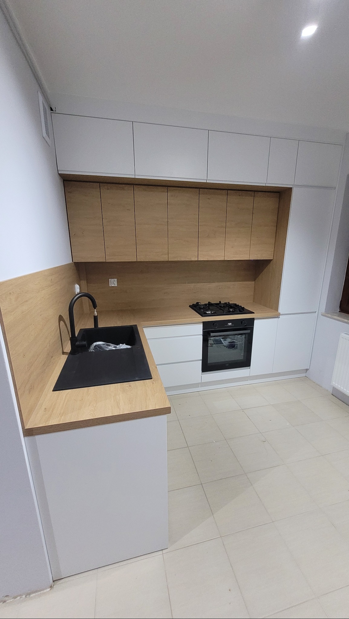 Nowoczesna kuchnia w Nowych Kozłowicach po remoncie: białe szafki dolne i górne, drewnopodobny blat i panel ścienny, czarny zlew i bateria, płyta gazowa i piekarnik.