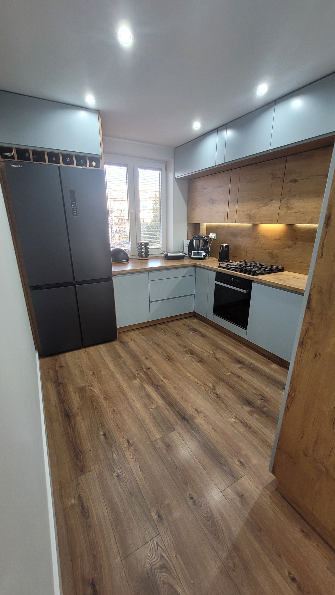 Nowoczesna kuchnia z drewnianymi panelami na podłodze i szafkami w kolorze pastelowej szarości, widoczna czarna, czterodrzwiowa lodówka Toshiba i drewnopodobne panele ścienne