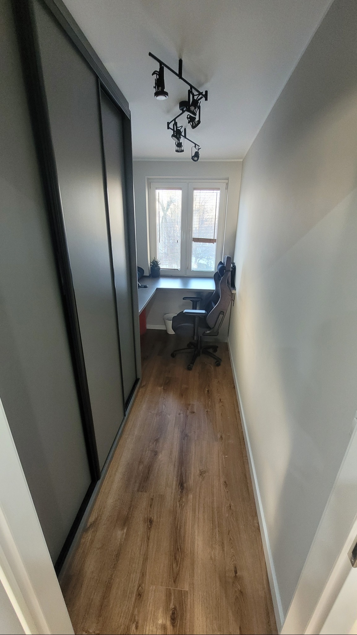 Wąskie pomieszczenie z drewnianą podłogą, biurkiem pod oknem i szarą szafą przesuwną. Czarna lampa sufitowa z kilkoma reflektorami oświetla przestrzeń.