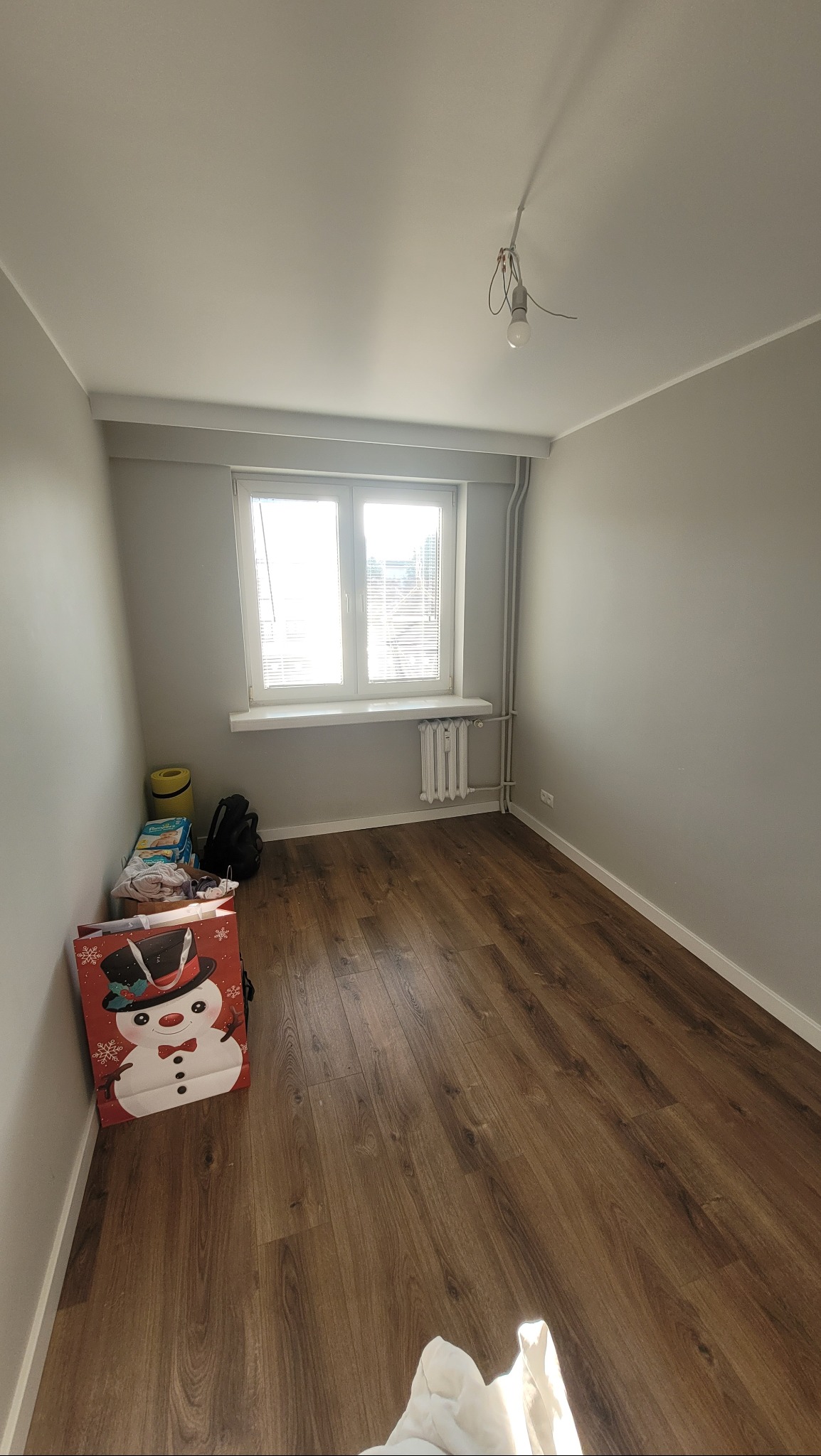 Pokój z brązowymi panelami podłogowymi, szarymi ścianami, grzejnikiem pod oknem i wiszącą żarówką bez klosza, torba prezentowa ze śnieżynką przy ścianie.