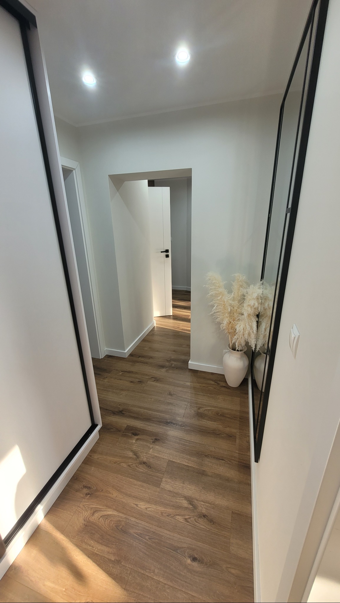Nowoczesny korytarz po remoncie z panelami podłogowymi w odcieniach brązu, białymi ścianami, czarną ramą lustra i dekoracyjnym wazonem z suszonymi trawami.