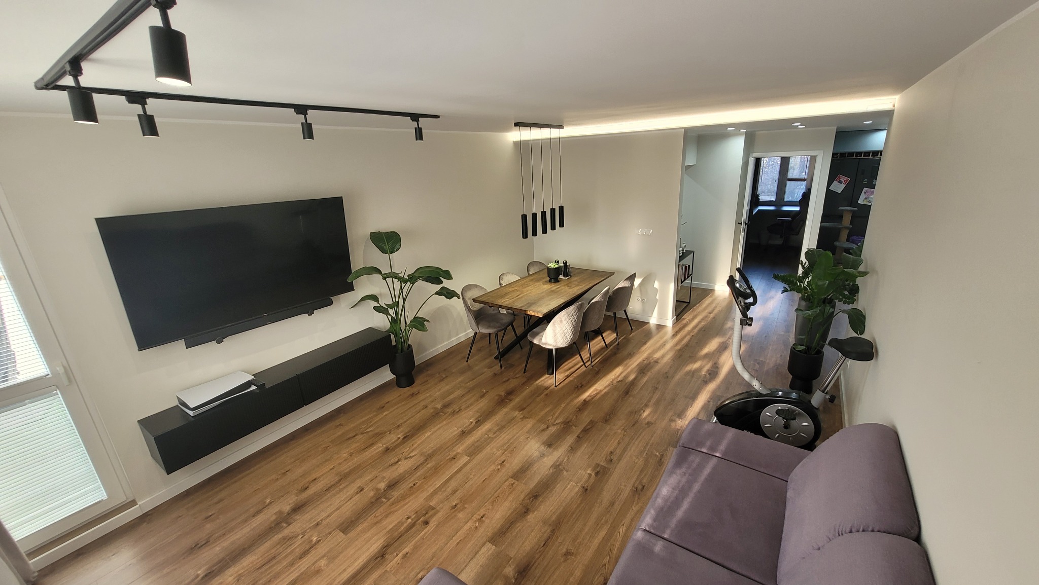 Jasne, przestronne wnętrze mieszkania z drewnianą podłogą, dużym telewizorem na ścianie, stołem z krzesłami i roślinami doniczkowymi, oświetlone nowoczesnymi lampami sufitowymi.