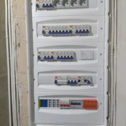 Rozdzielnica elektryczna z zabezpieczeniami i gniazdami, widoczne przewody w peszlach. Biała obudowa na surowej ścianie.