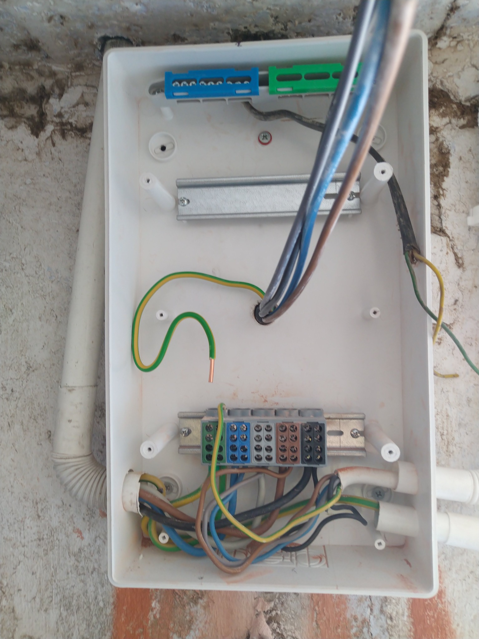 Otwarta, biała skrzynka elektryczna z widocznymi przewodami różnej grubości i koloru, złączkami i listwami montażowymi. Detal instalacji na tle ściany.