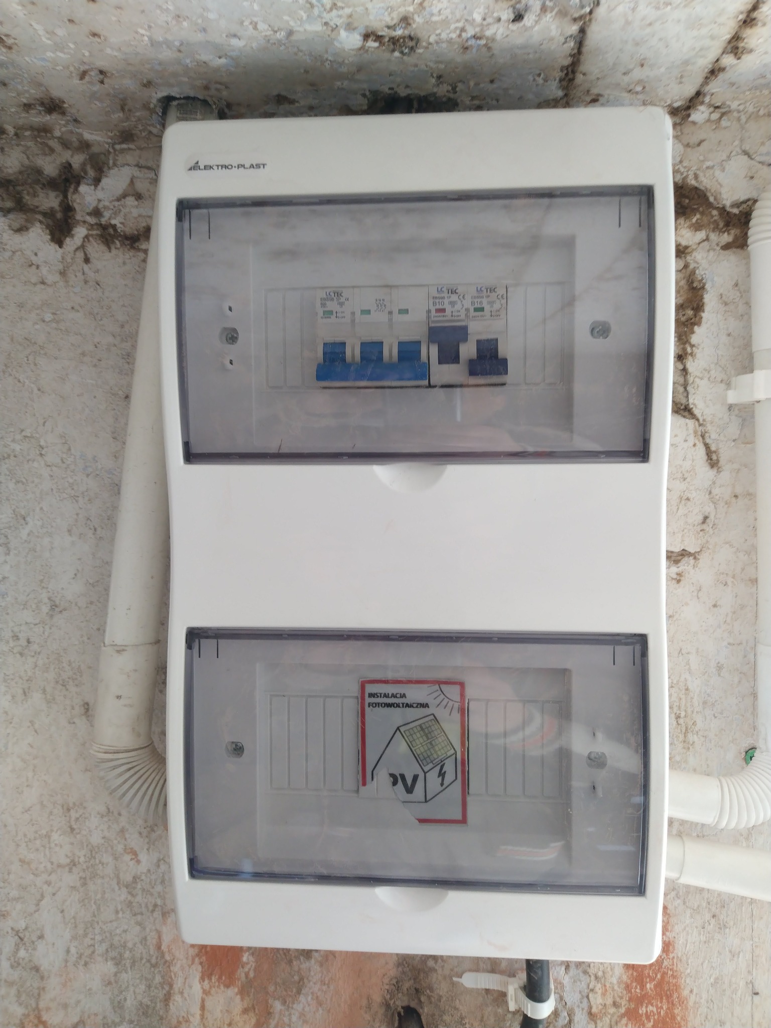 Biała skrzynka elektryczna Elektro-Plast z bezpiecznikami i naklejką 'Instalacja Fotowoltaiczna PV' na tle ściany z widoczną instalacją.
