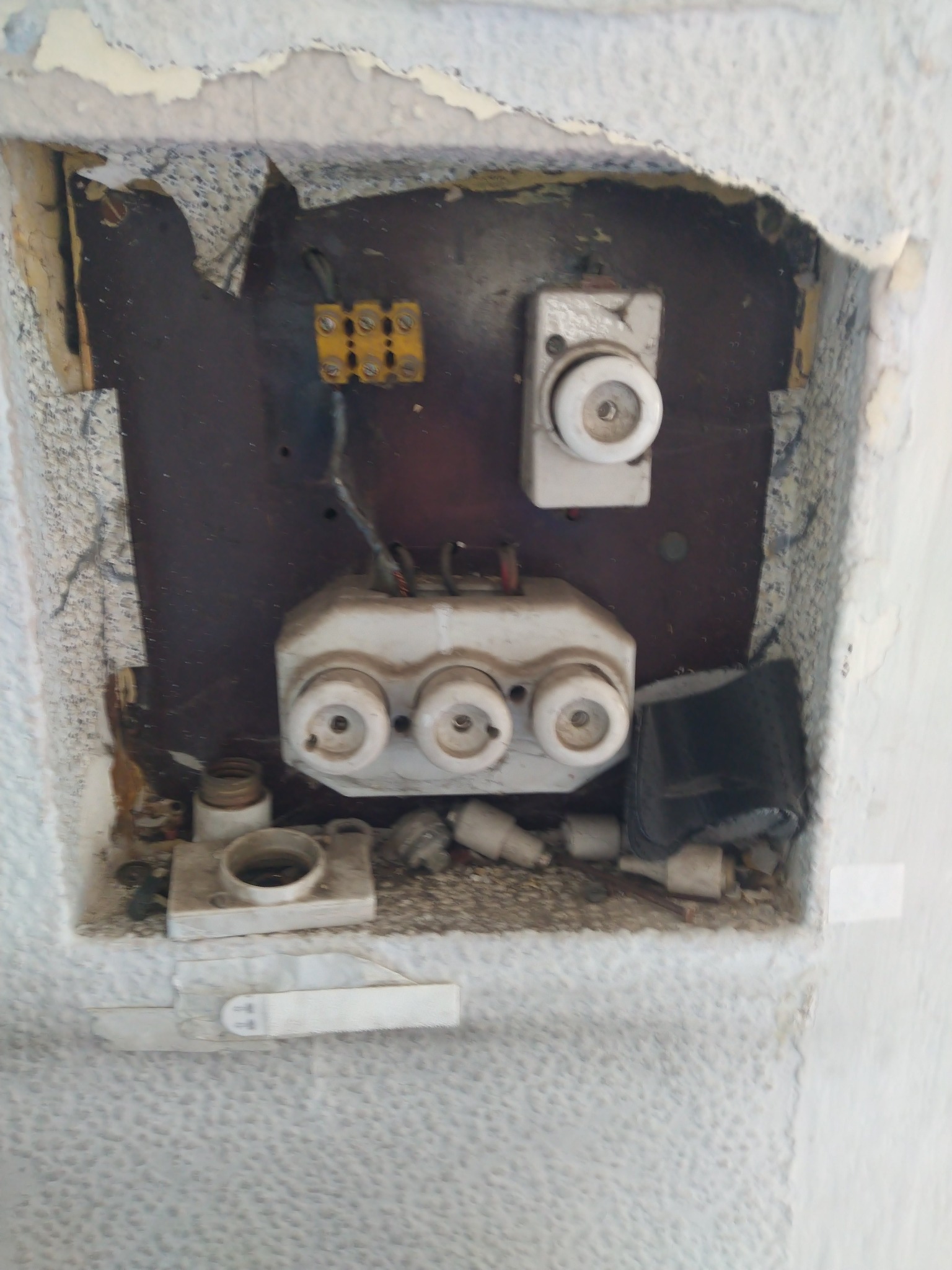 Stara tablica bezpiecznikowa z ceramicznymi wkładkami, widoczna w ścianie z odspojoną farbą i fragmentami tapety, ukazująca zużycie i potencjalną potrzebę modernizacji.