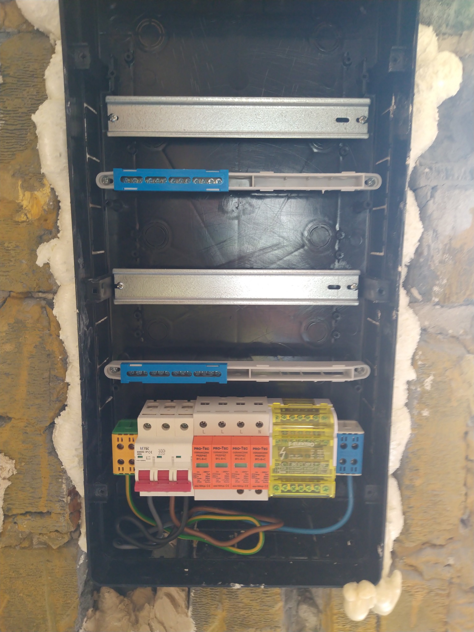 Otwarta skrzynka elektryczna z zamontowanymi zabezpieczeniami przepięciowymi i przewodami, widoczna na ścianie z pianką montażową.