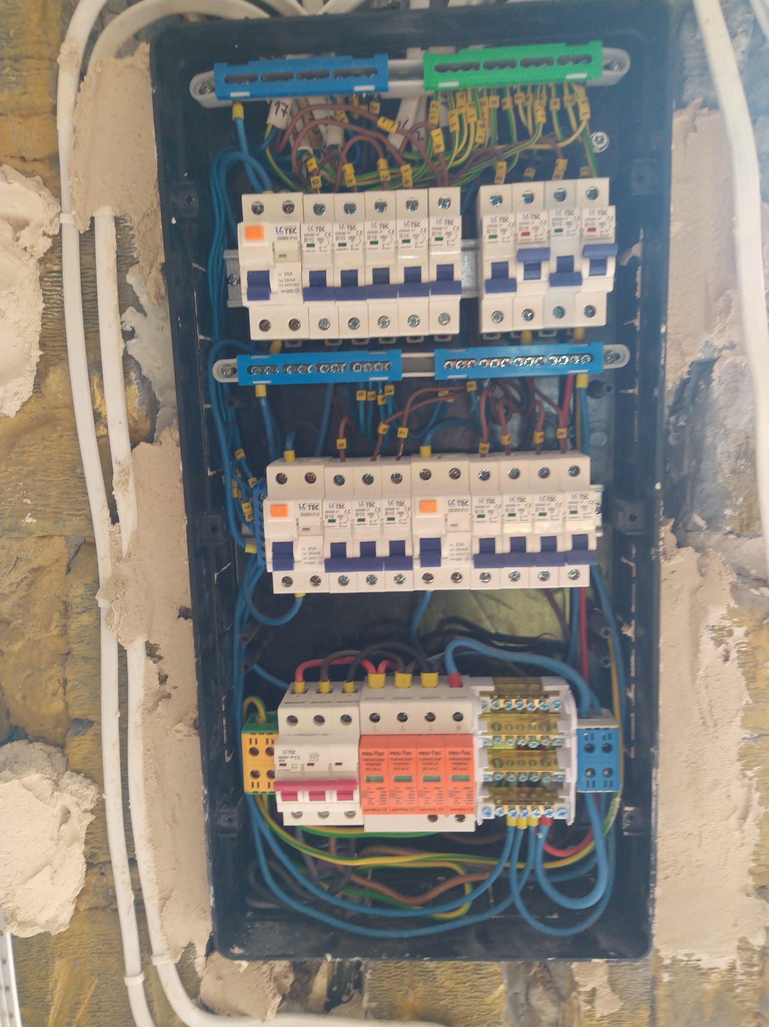 Rozdzielnica elektryczna z bezpiecznikami i przewodami w trakcie montażu, widoczne elementy instalacji na ścianie w surowym stanie.