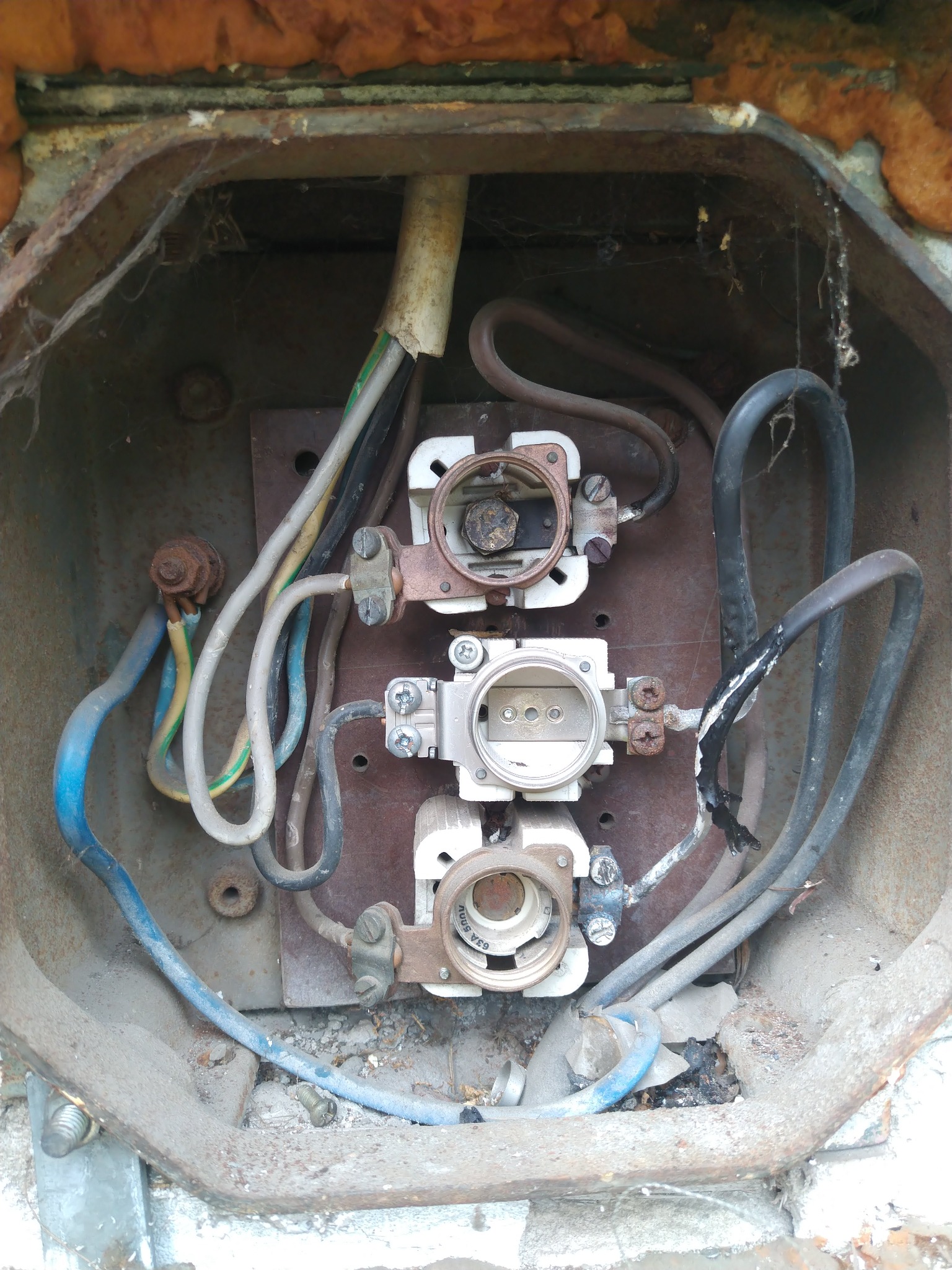 Stara, skorodowana skrzynka bezpiecznikowa z widocznymi przewodami, bezpiecznikami i śladami korozji. Widoczne pajęczyny i zabrudzenia wskazują na zaniedbanie.