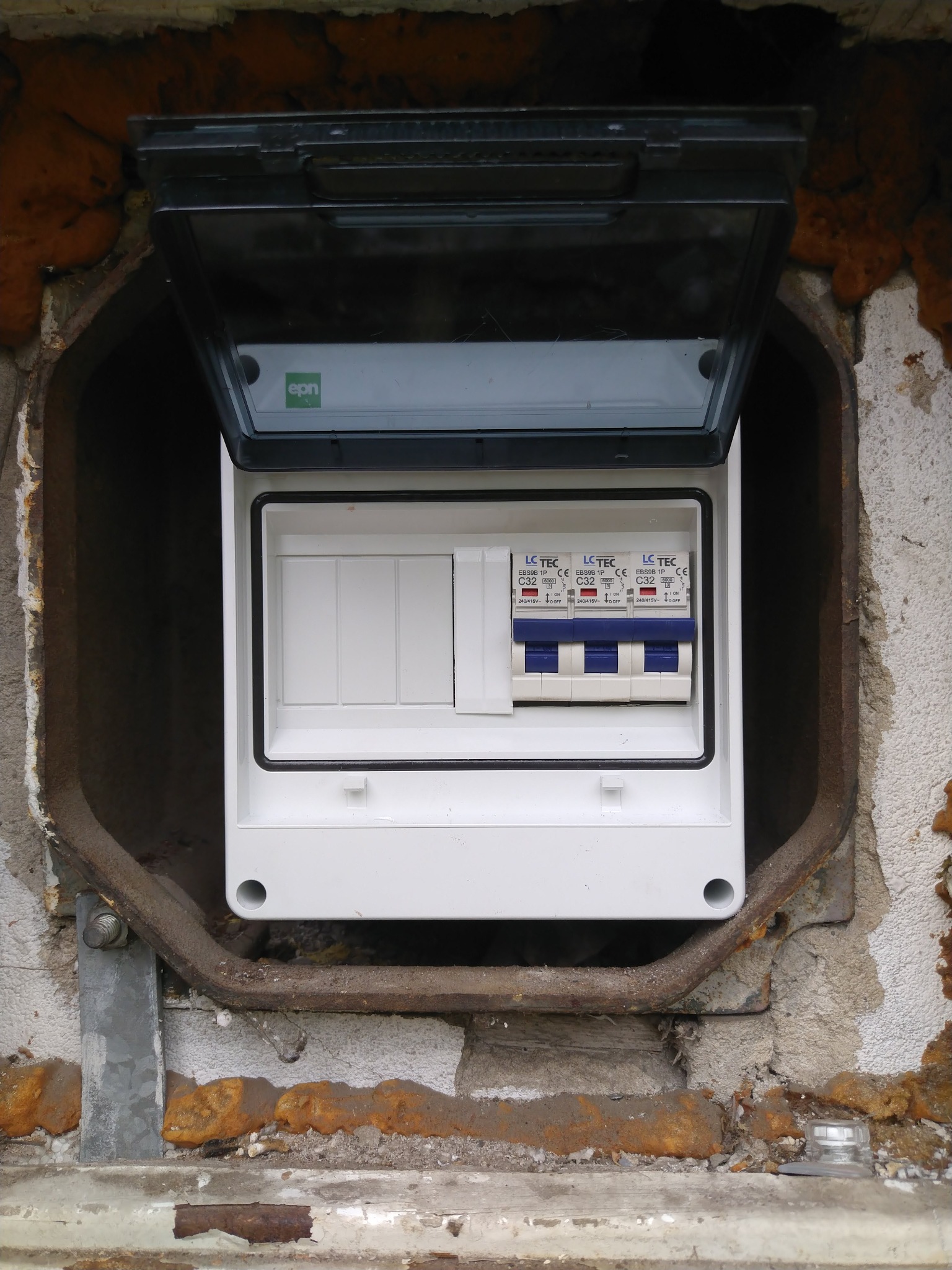 Otwarta, biała skrzynka elektryczna z bezpiecznikami LC TEC, zamontowana w ścianie z widocznymi śladami prac instalacyjnych i pianką montażową.