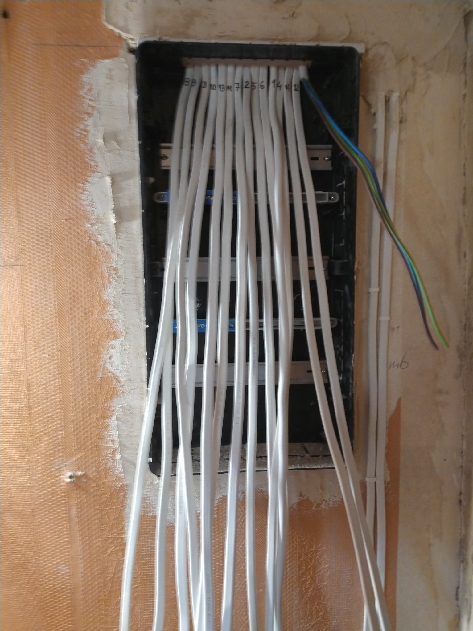 Rozprowadzone białe przewody w czarnej skrzynce instalacyjnej w ścianie, z numeracją. Widoczne kable w kolorowej izolacji. Ściana w trakcie prac wykończeniowych.
