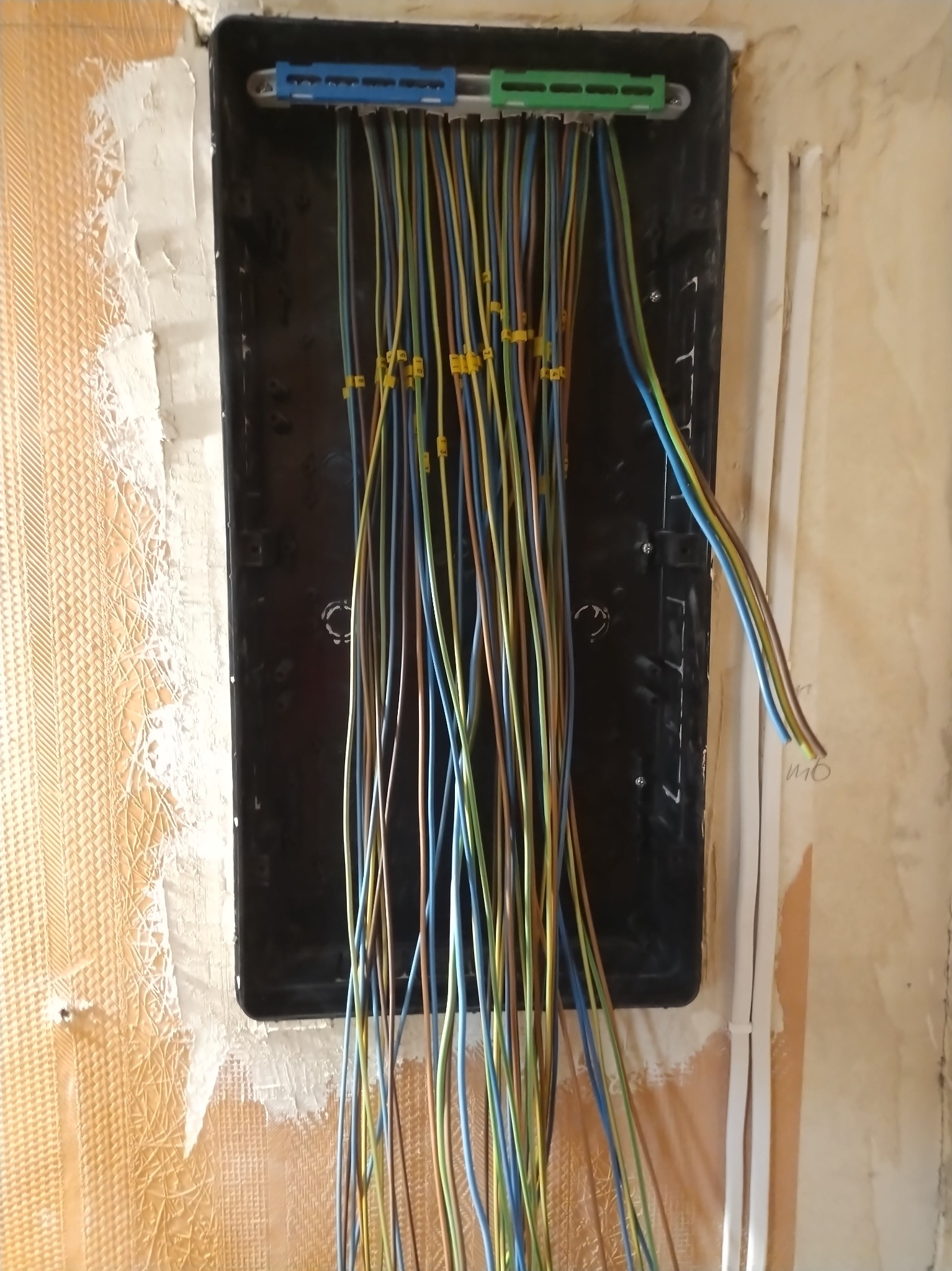 Czarna skrzynka elektryczna z podłączonymi przewodami o różnych kolorach, zamontowana na ścianie z widocznymi śladami po remoncie. Widoczne żółte oznaczenia na kablach.