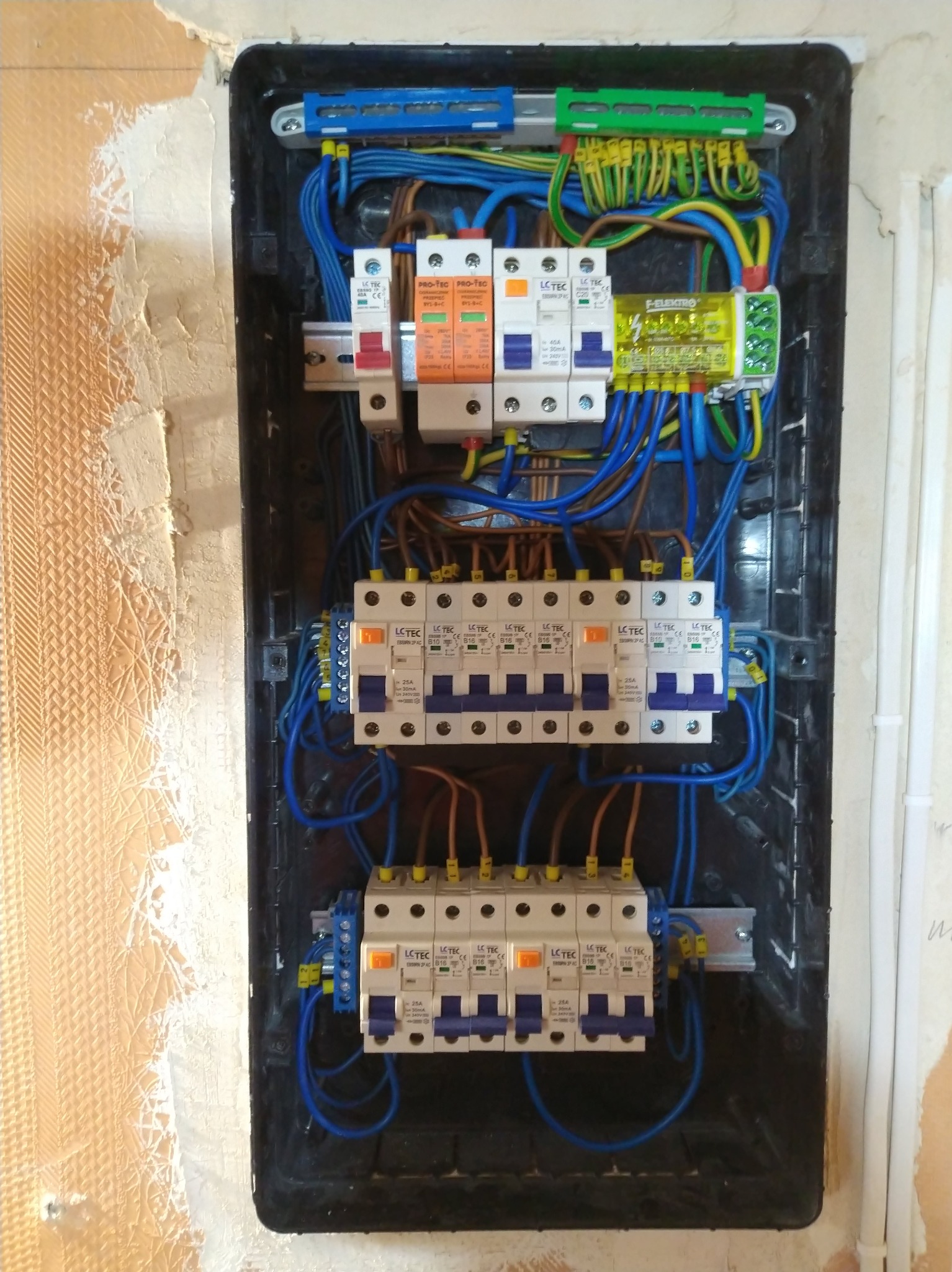 Rozdzielnica elektryczna w czarnej obudowie, widoczne liczne kable, bezpieczniki i listwy zaciskowe. Ściana wokół w trakcie wykończenia, widoczne ślady po gładzi.