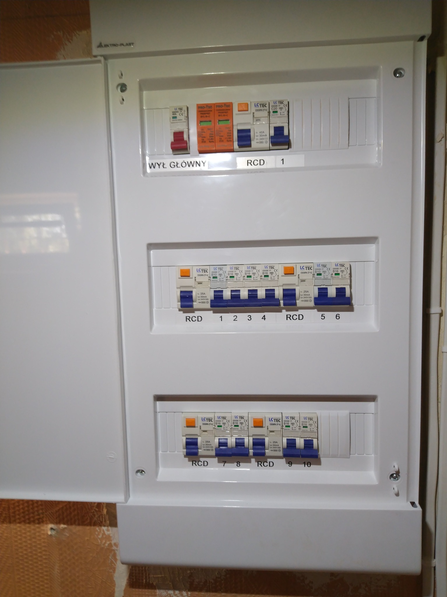 Otwarta skrzynka elektryczna z bezpiecznikami, widoczne wyłączniki RCD i oznaczenia prądowe, zamontowana na ścianie z widoczną fakturą. Oświetlenie górne.