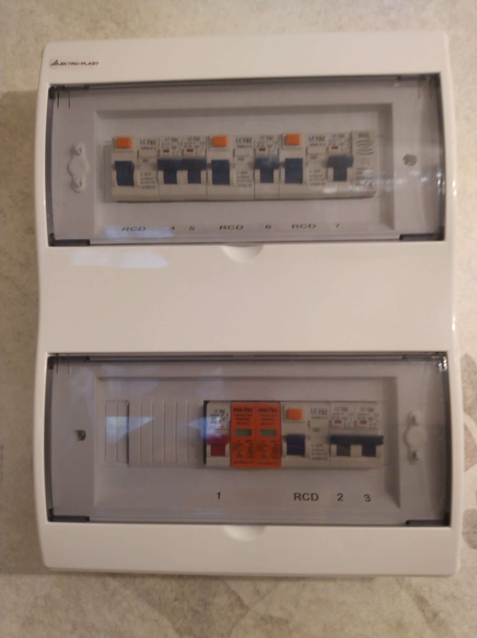 Dwudzielna, biała skrzynka elektryczna z widocznymi bezpiecznikami i oznaczeniami RCD, zamontowana na ścianie o delikatnej strukturze. Widoczne logo Elektro-Plast.