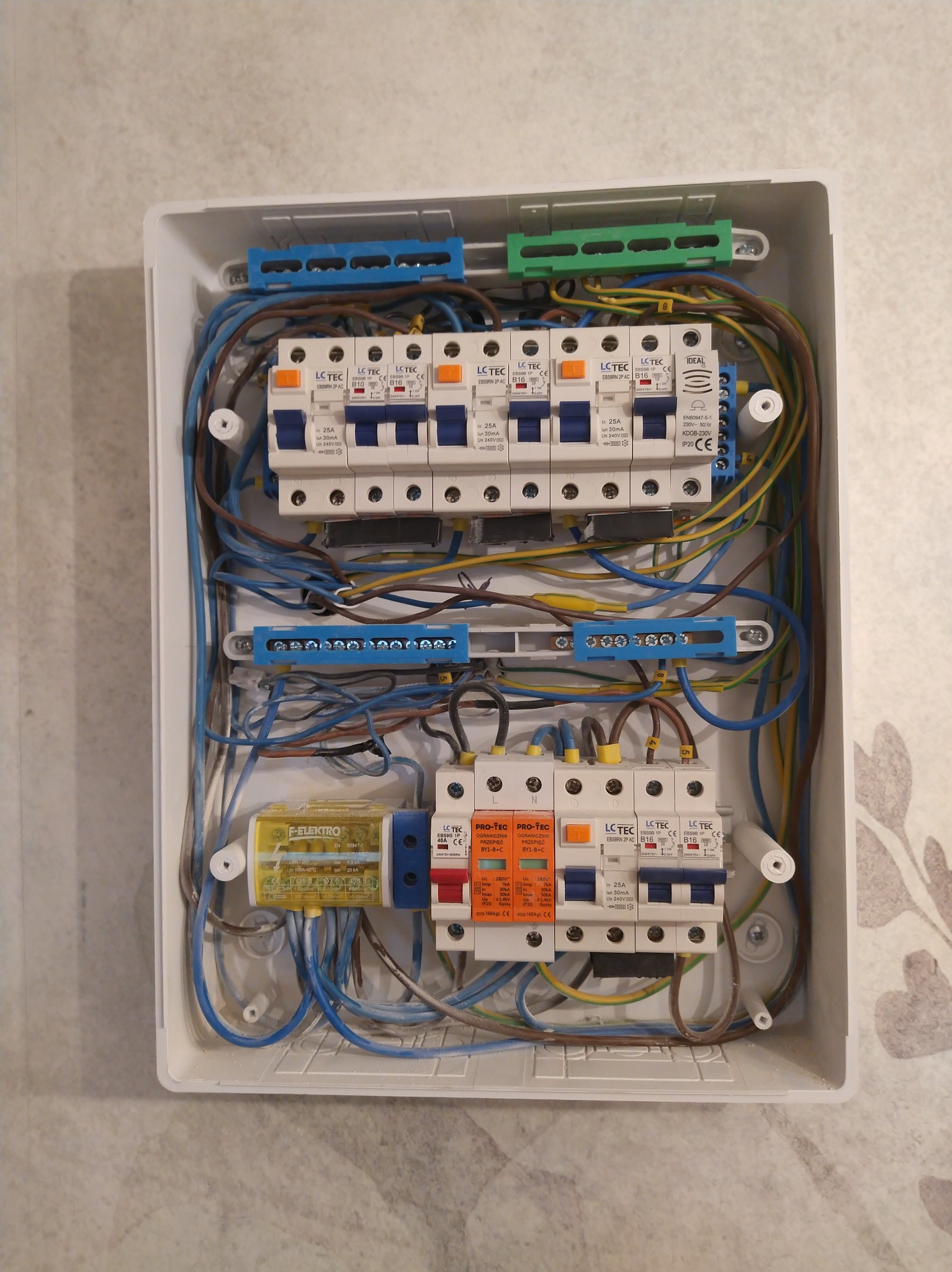 Otwarta skrzynka elektryczna z zamontowanymi bezpiecznikami, widoczne przewody w różnych kolorach, elementy montażowe i naklejki informacyjne.
