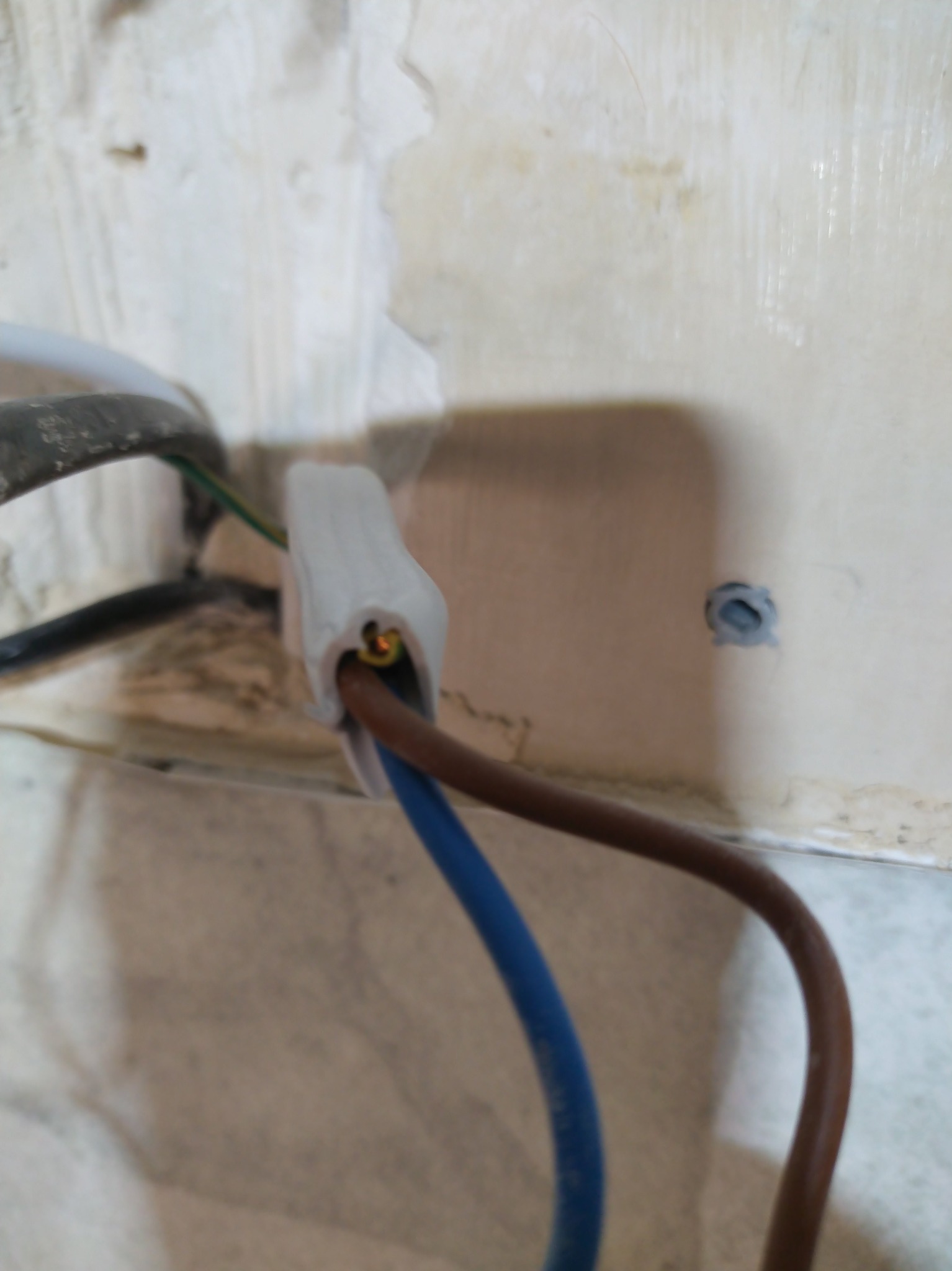 Złącze elektryczne z przewodami (niebieski, brązowy) na tle ściany z widocznymi śladami po instalacji. Widoczne kable w izolacji i fragment ściany z otworem.