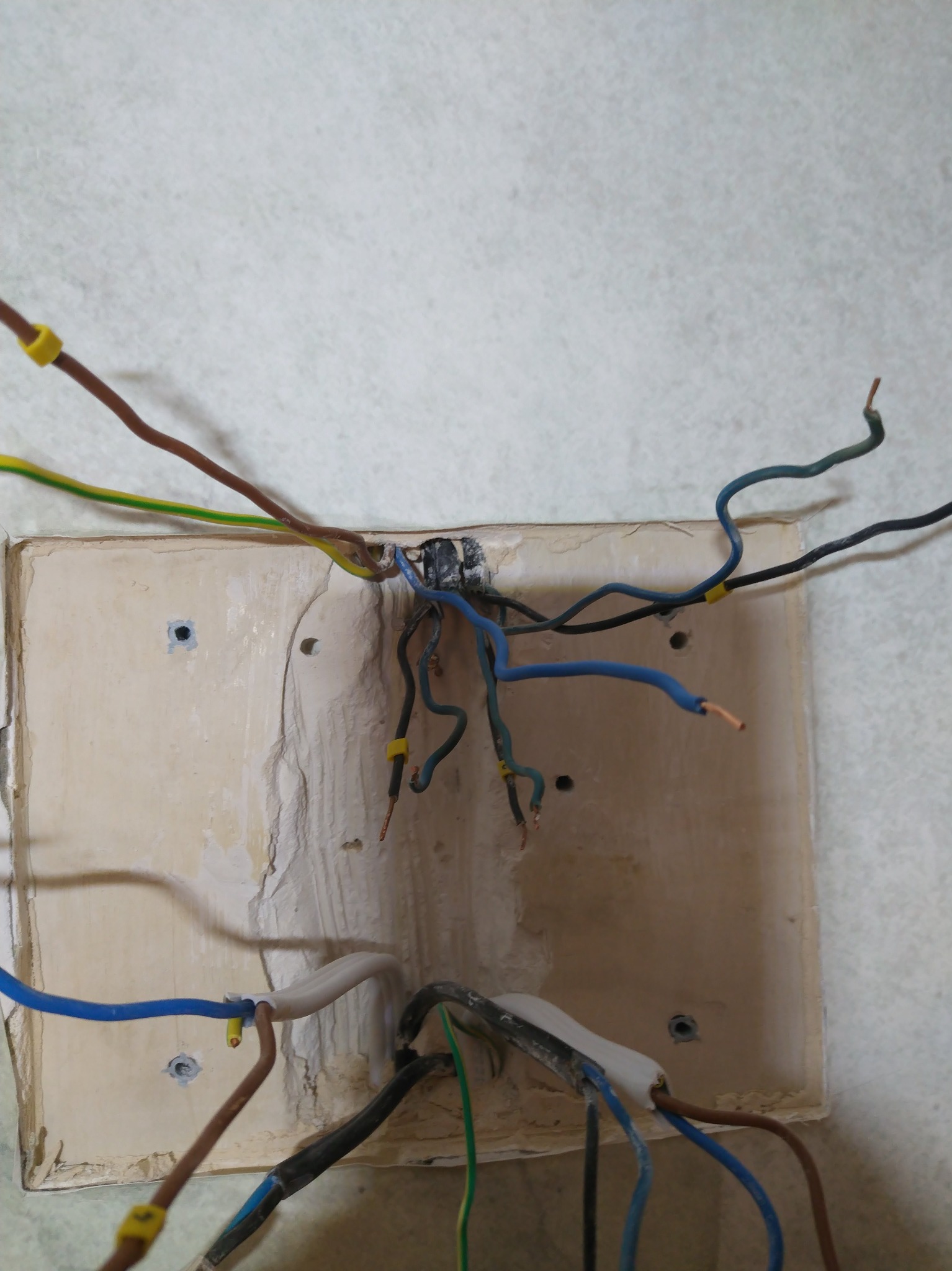Niezabezpieczone, wystające przewody elektryczne z żółtymi oznaczeniami, wychodzące ze ściany. Widoczne różne kolory kabli oraz fragment puszki elektrycznej.