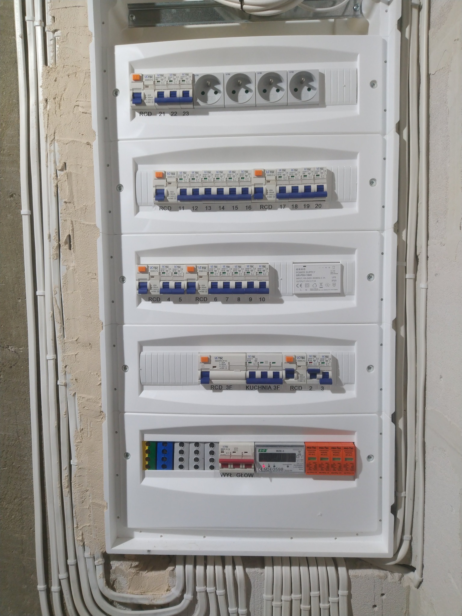 Rozdzielnica elektryczna z zabezpieczeniami i gniazdami, widoczne przewody w peszlach. Biała obudowa na surowej ścianie.