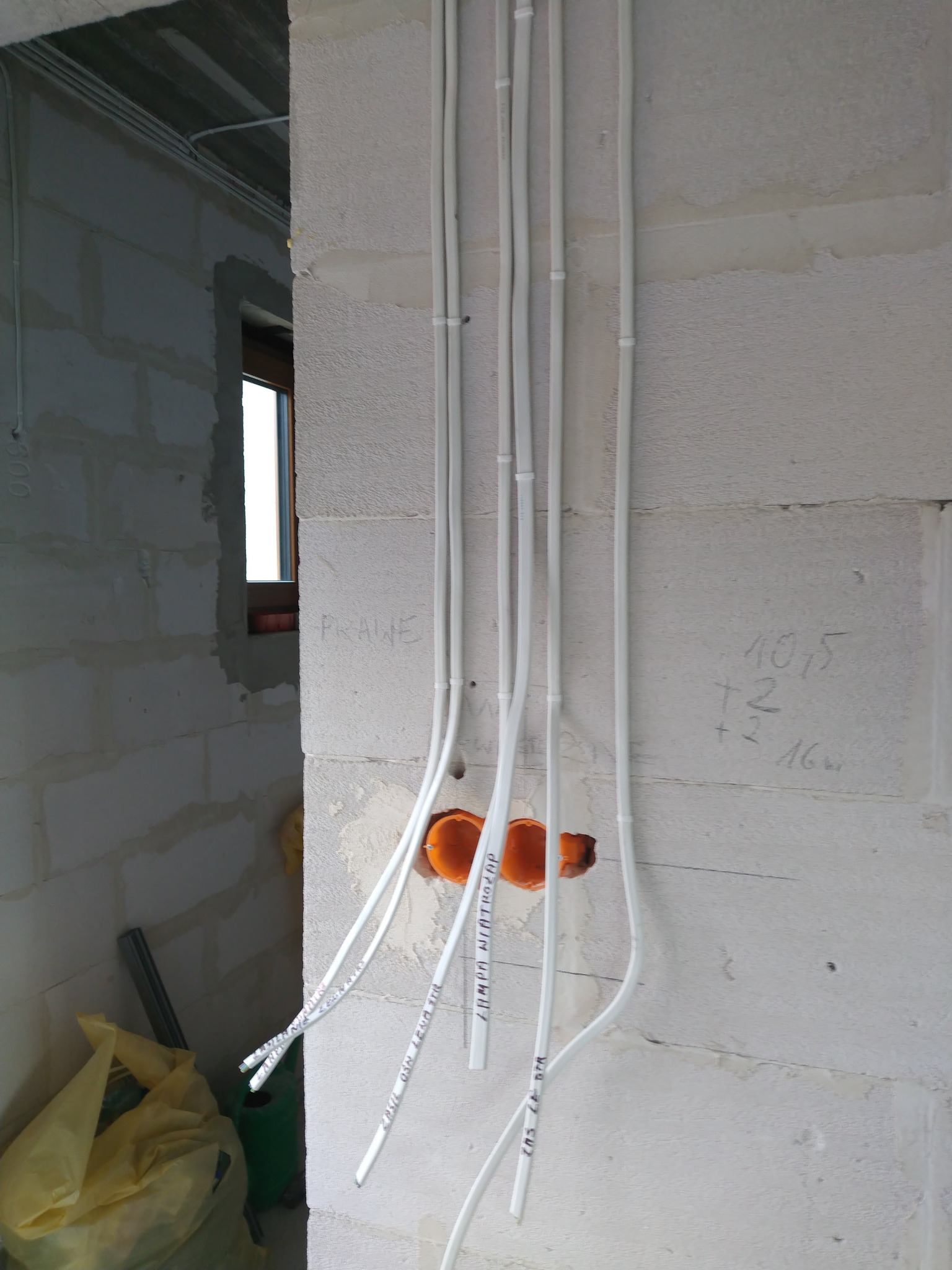 Instalacja elektryczna w surowym wnętrzu: białe kable prowadzone po ścianie z bloczków, widoczne puszki instalacyjne i napisy na kablach. W tle okno i surowa ściana.