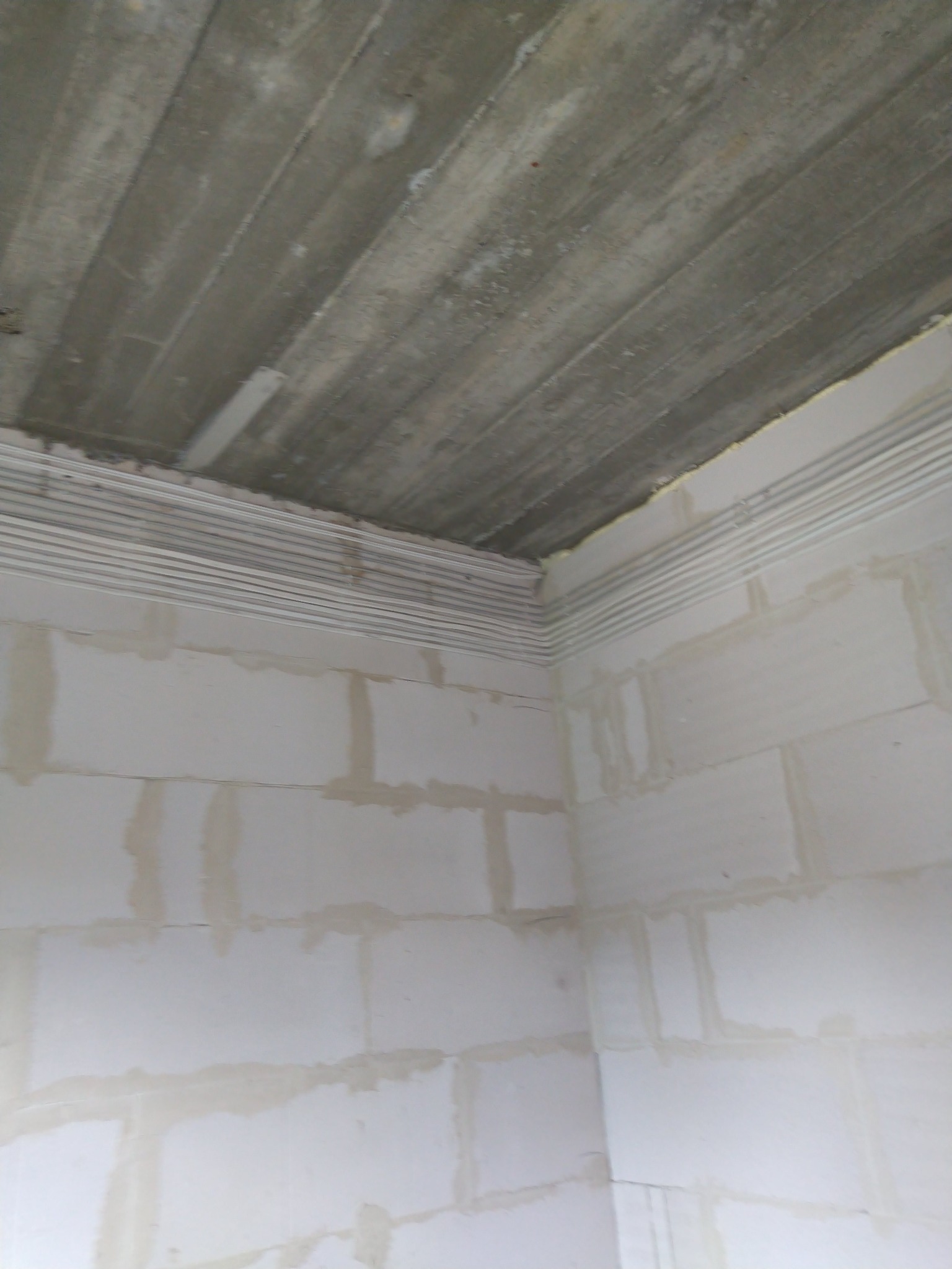 Surowy betonowy strop z widocznymi deskami szalunkowymi i ściany z białych bloczków z szarymi fugami, przygotowane do dalszych prac wykończeniowych.