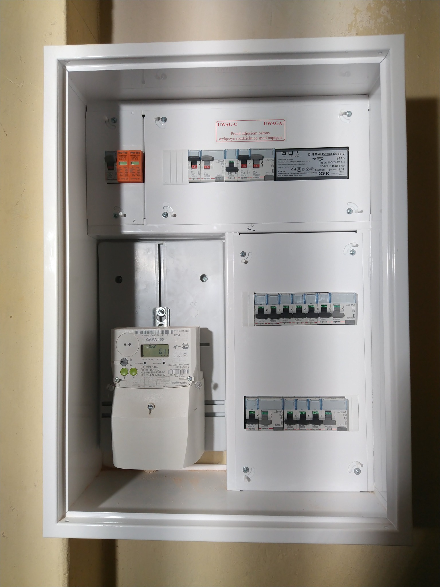 Biała skrzynka elektryczna z licznikiem i zabezpieczeniami, zamontowana na ścianie. Widoczne bezpieczniki, wyłączniki różnicowoprądowe i tabliczka znamionowa z ostrzeżeniem.