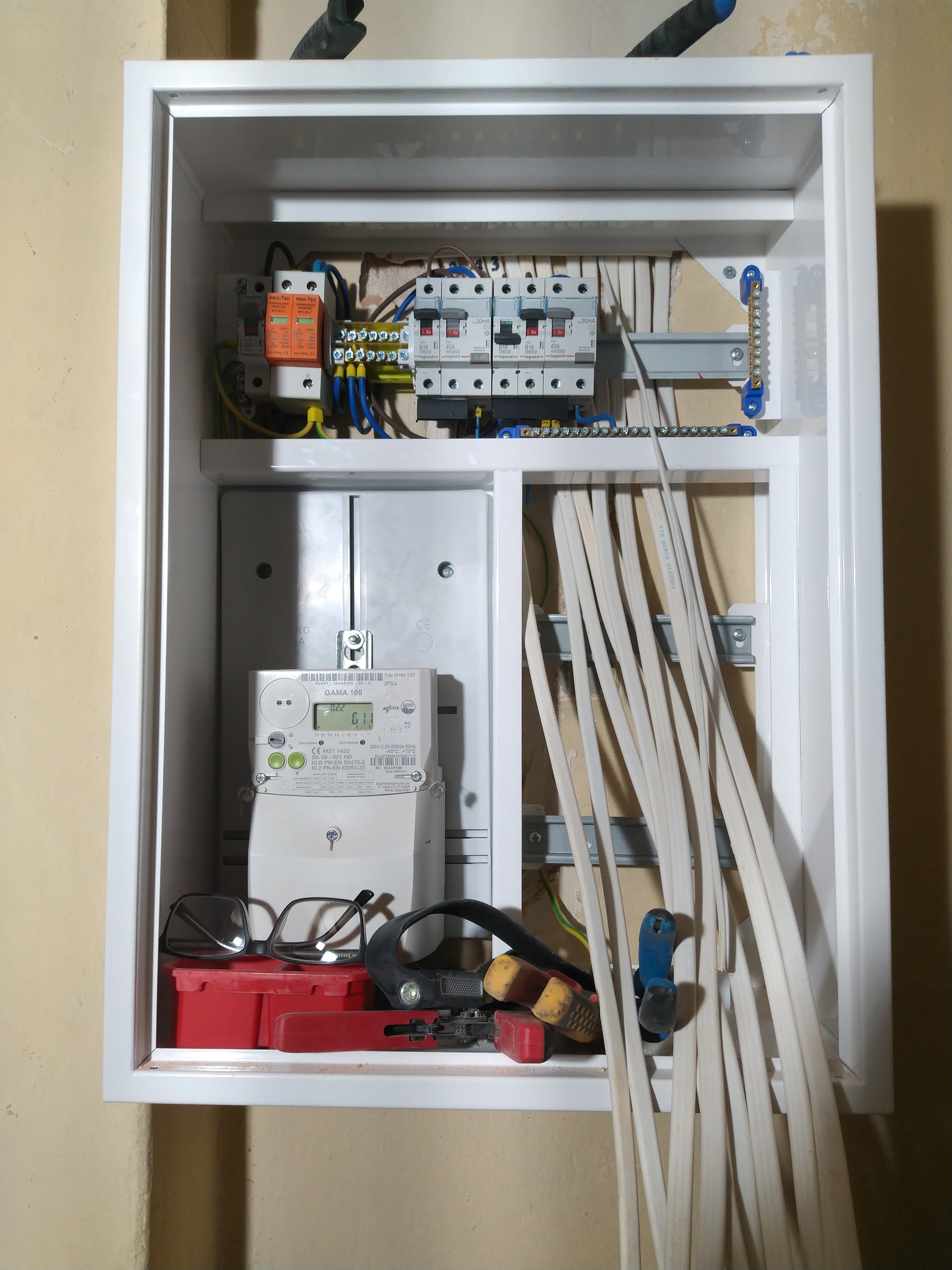 Rozdzielnica elektryczna w białej obudowie, widoczne zabezpieczenia, przewody, licznik energii, narzędzia i okulary na dole. Ujęcie z bliska, oświetlenie naturalne.