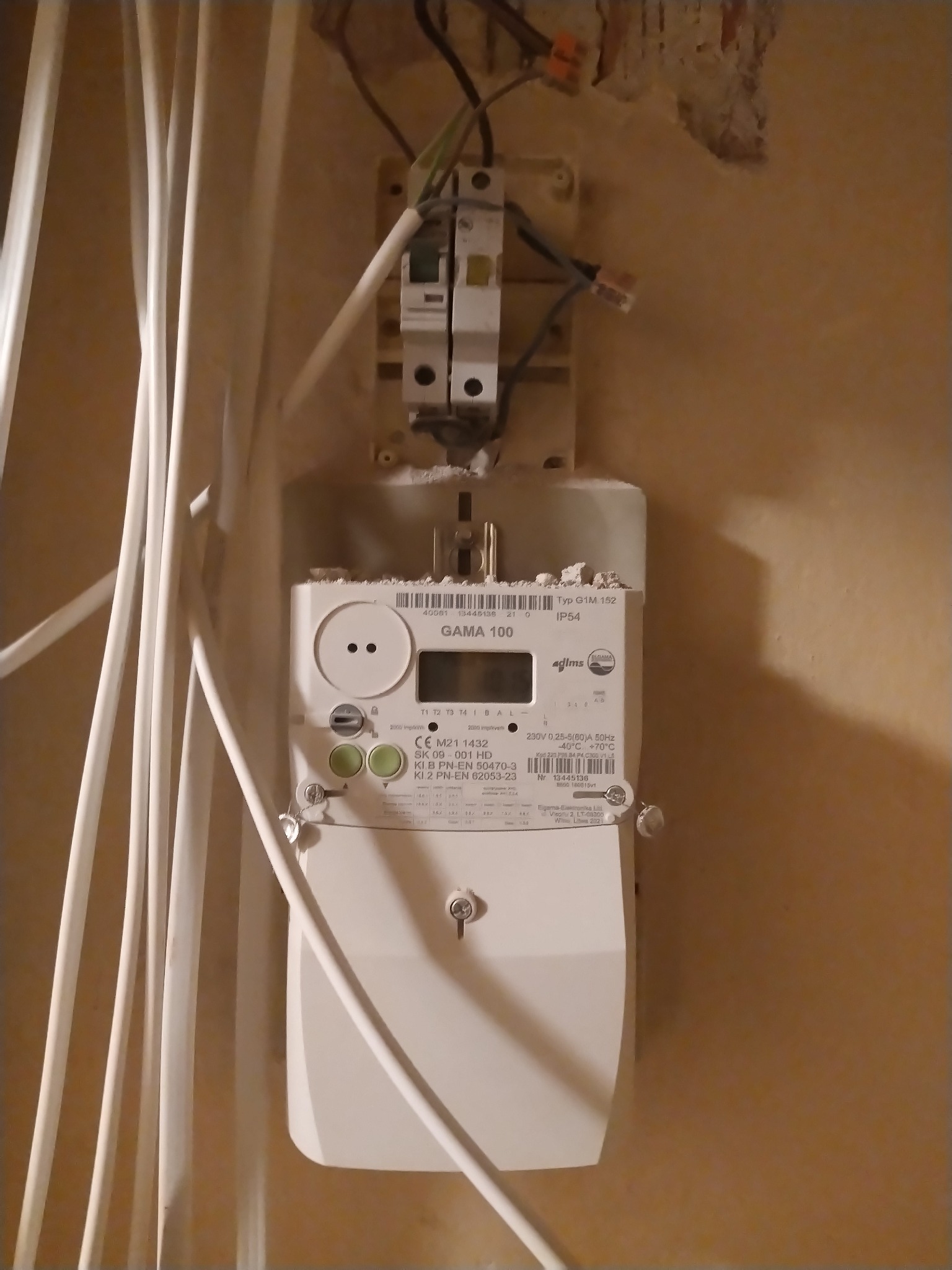 Zamontowany licznik elektryczny Gama 100 na ścianie z widocznymi przewodami i zabezpieczeniami. Widoczne ślady po kuciu w ścianie.
