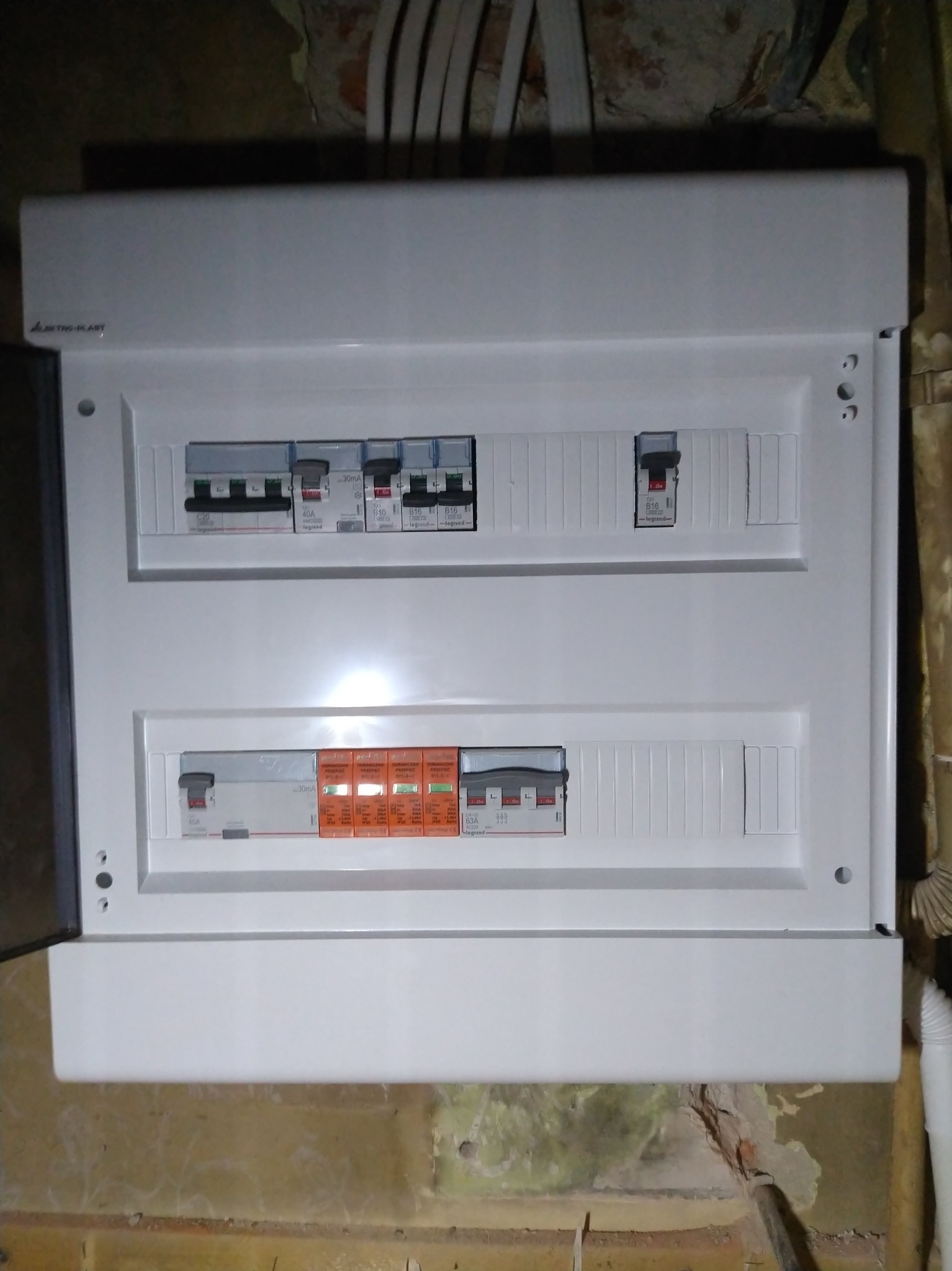 Biała skrzynka rozdzielcza z bezpiecznikami i ogranicznikiem przepięć, zamontowana na ścianie z widocznymi śladami po instalacji. Otwarta obudowa po lewej stronie.