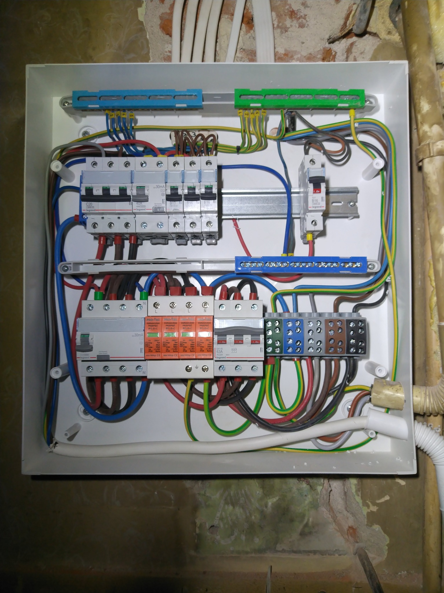 Rozdzielnica elektryczna z zainstalowanymi bezpiecznikami, ogranicznikami przepięć i kolorowymi przewodami w białej obudowie, zamontowana na ścianie z widocznymi śladami prac murarskich.