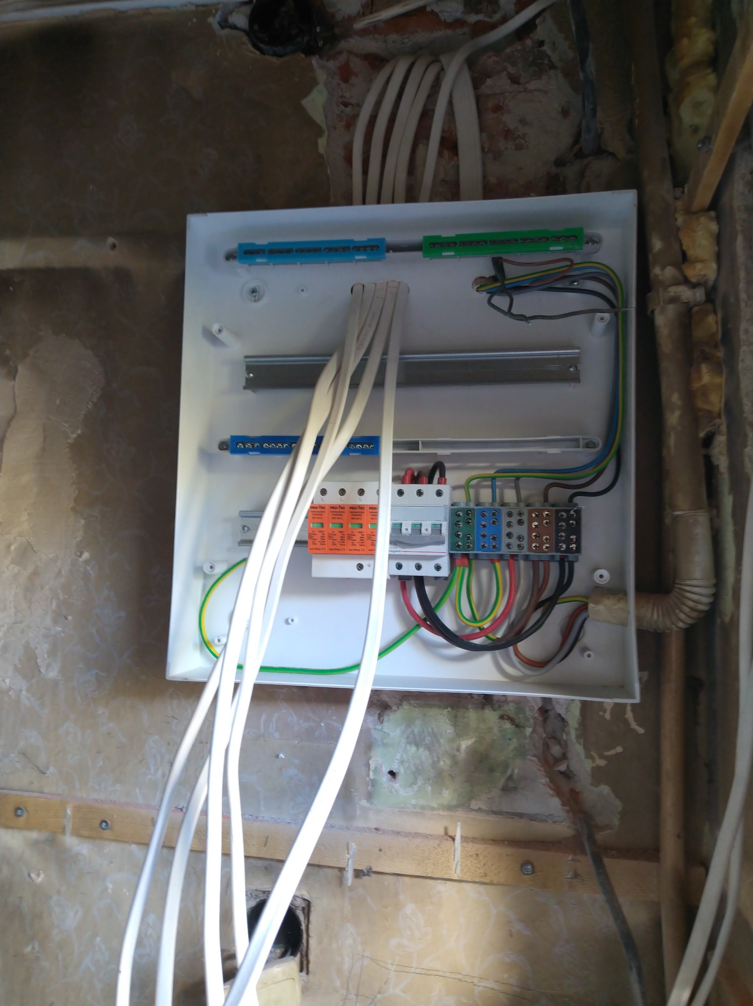 Rozdzielnica elektryczna w trakcie montażu na ścianie z widocznymi przewodami, elementami zabezpieczeń i listwami zaciskowymi. Ściana w stanie surowym.