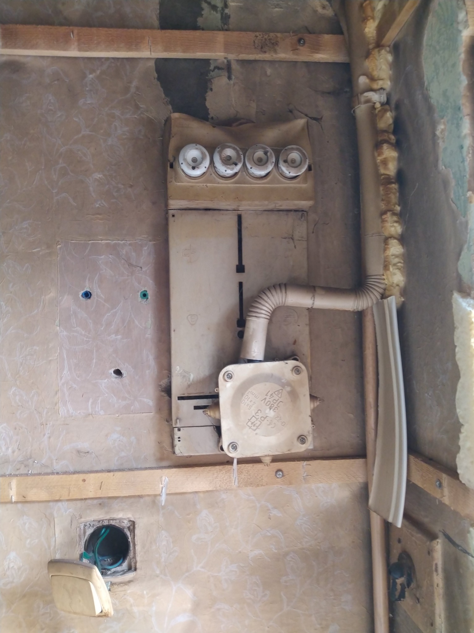 Stara skrzynka bezpiecznikowa z ceramicznymi wkładkami, rurą instalacyjną i puszką przyłączeniową na ścianie z tapetą w trakcie remontu. Widoczne drewniane elementy konstrukcyjne.