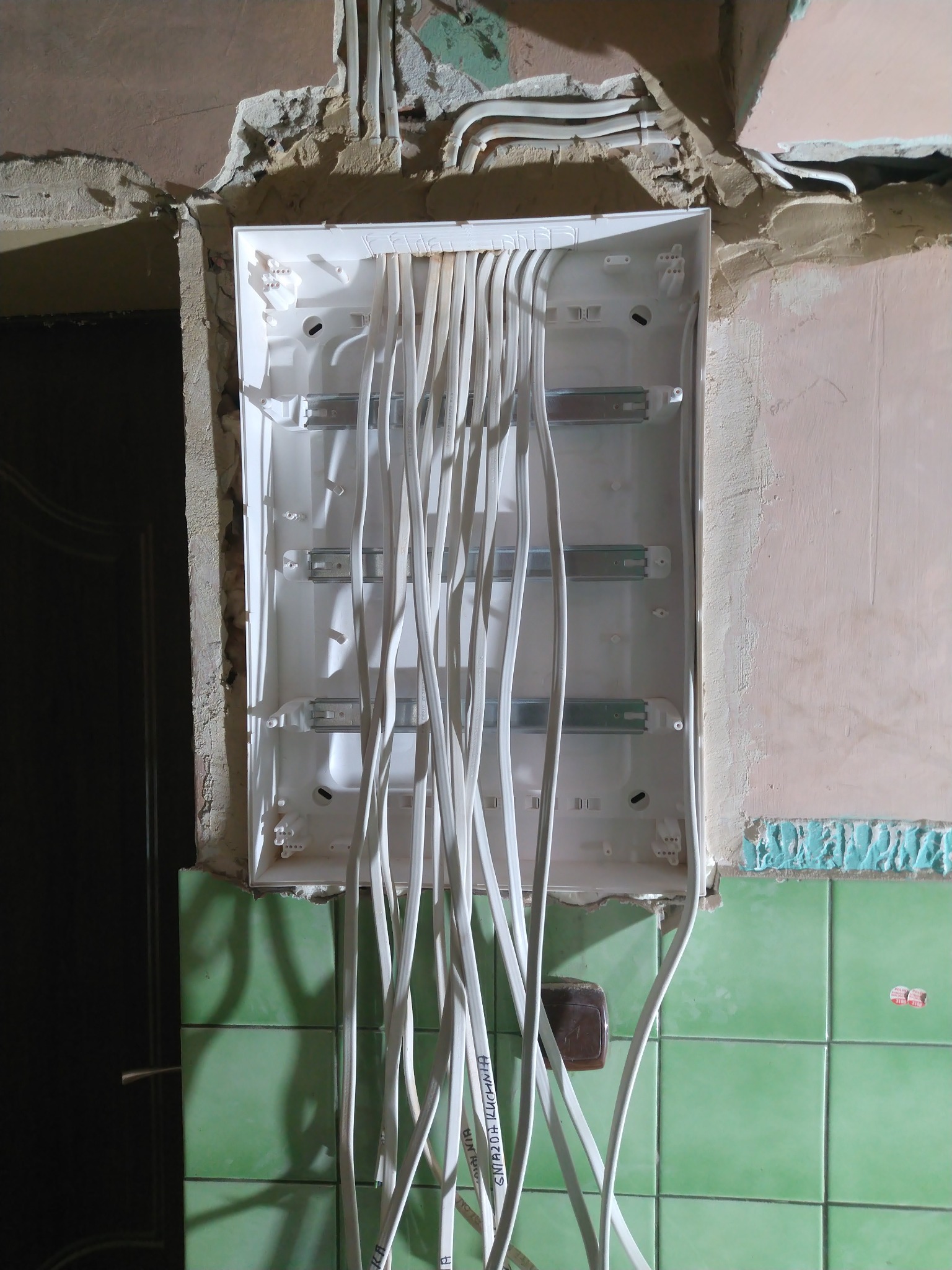 Biała skrzynka rozdzielcza z licznymi kablami w surowym wnętrzu. Widoczne ślady po kuciu w ścianie, zielone płytki i fragment drzwi. Etap prac elektrycznych.