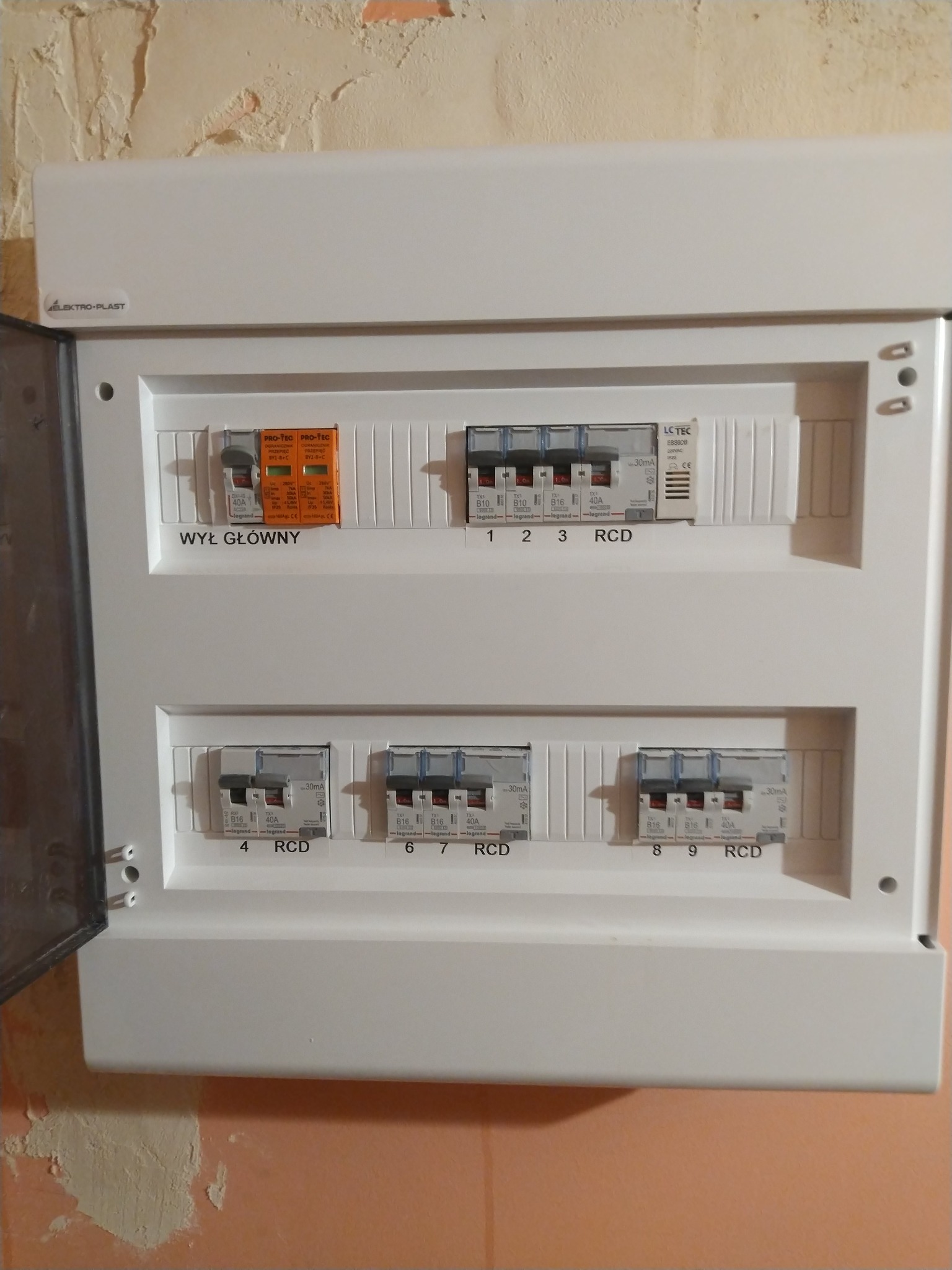 Rozdzielnica elektryczna Elektro-Plast z bezpiecznikami Legrand i LC TEC, widoczne oznaczenia RCD i wyłącznik główny, zamontowana na ścianie z surowym wykończeniem.