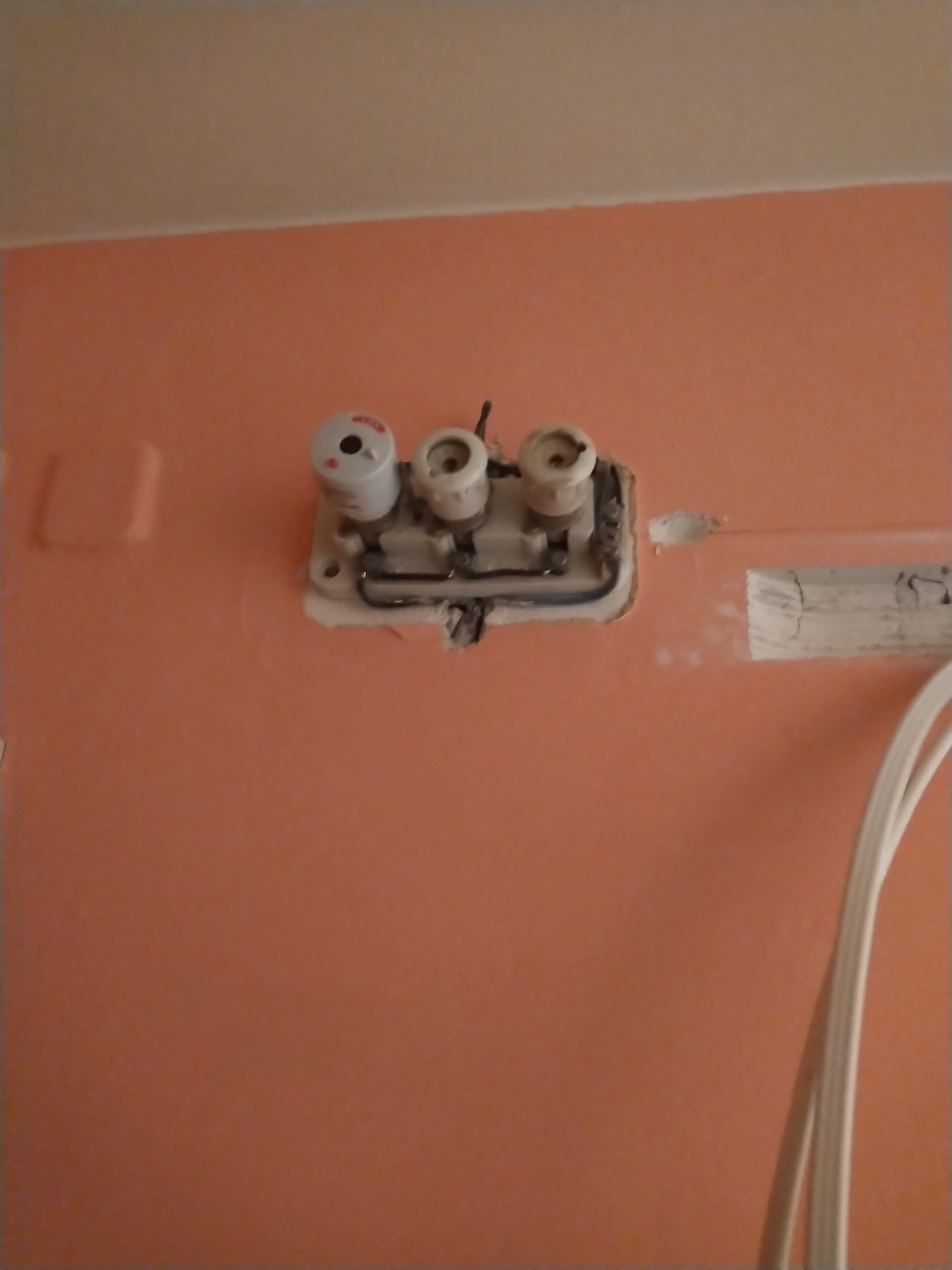 Gniazdo bezpiecznikowe z trzema bezpiecznikami na pomarańczowej ścianie, widoczne przewody. Drobne uszkodzenia ściany wokół gniazda.